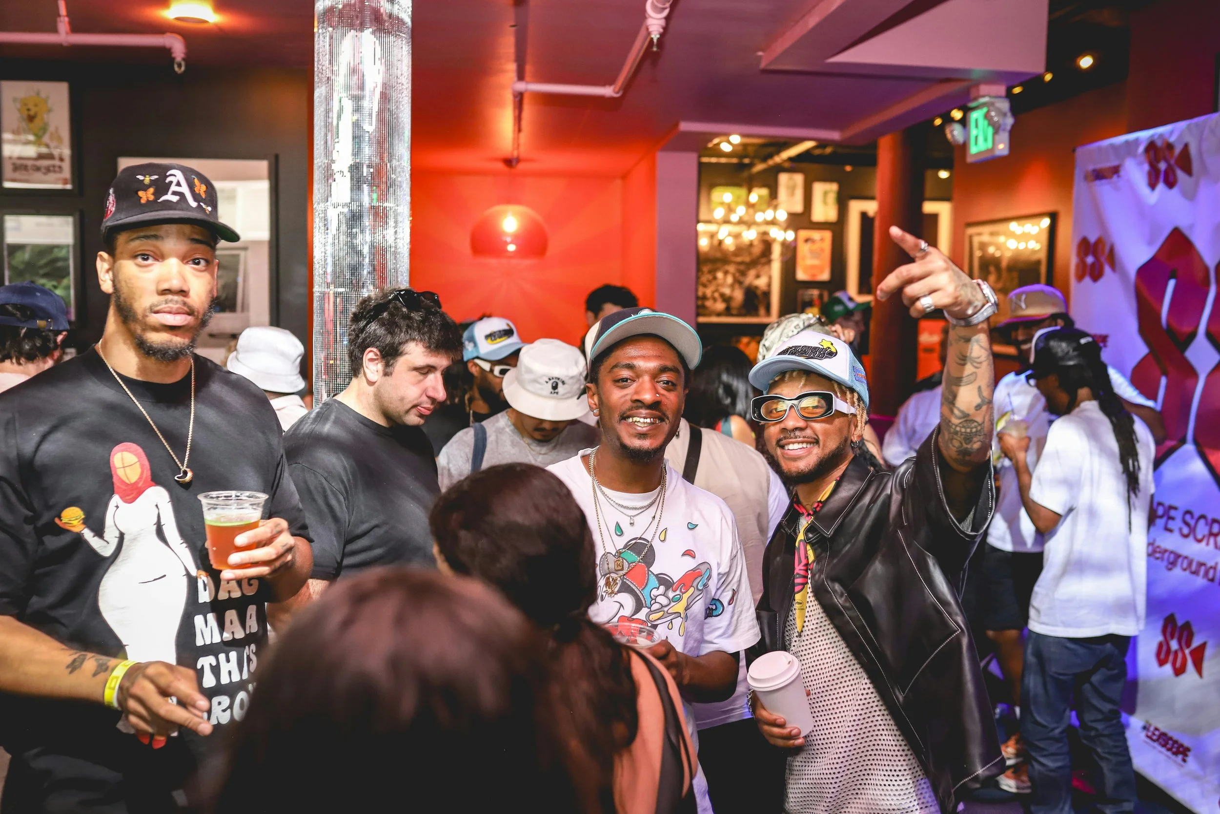 Group of people socializing at an indoor event or party with colorful lighting and wall decorations, some wearing hats and glasses, holding drinks.
