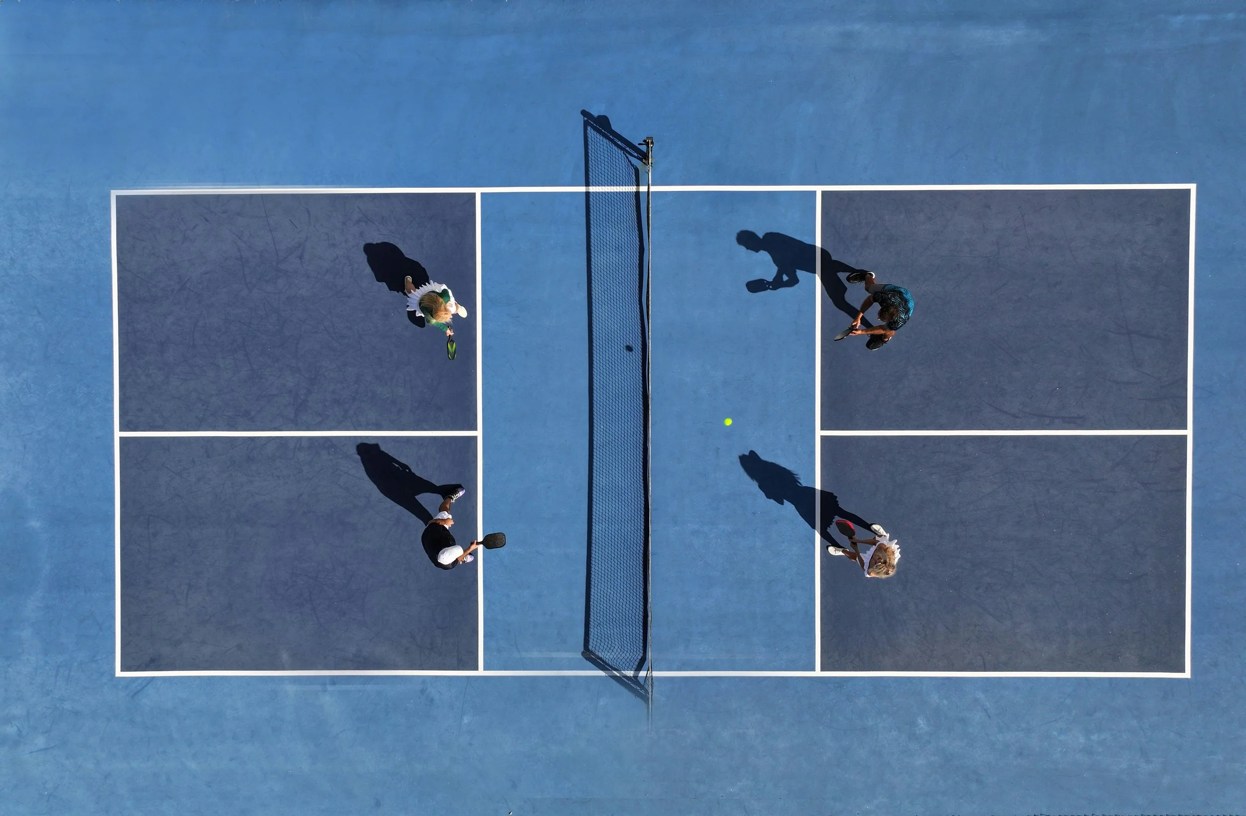 Four people playing pickleball on an outdoor court from above, with a green ball in play and shadows cast on the blue surface.