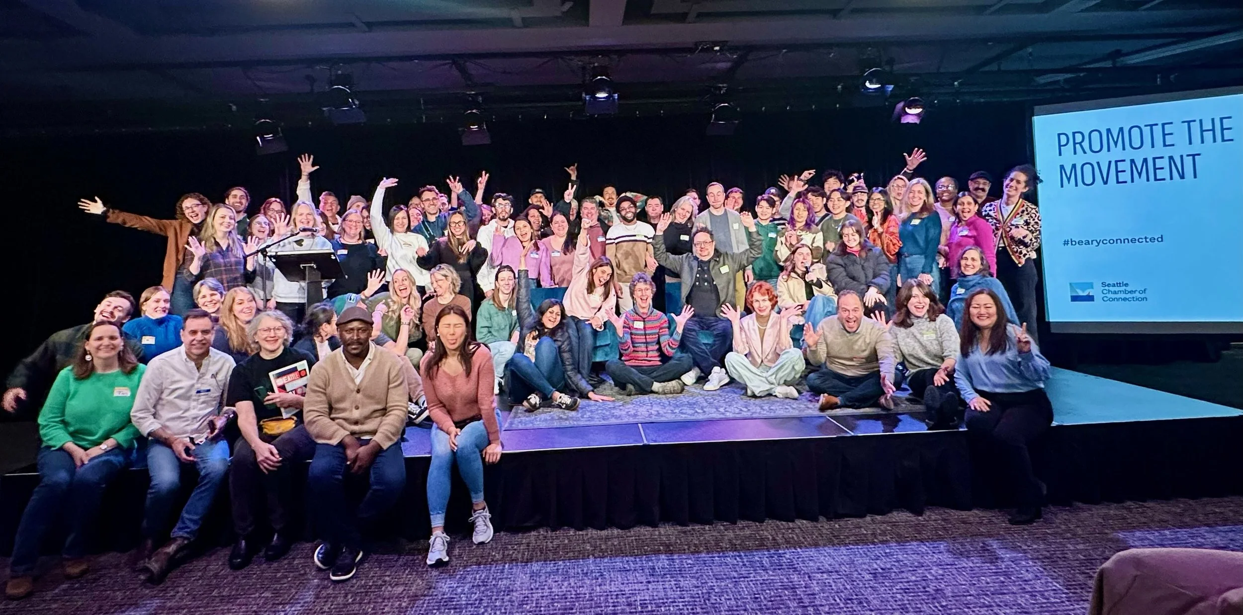Group of diverse people on stage at a conference, smiling and waving, with a screen displaying 'PROMOTE THE MOVEMENT' and '#bearcyconnected' in the background.