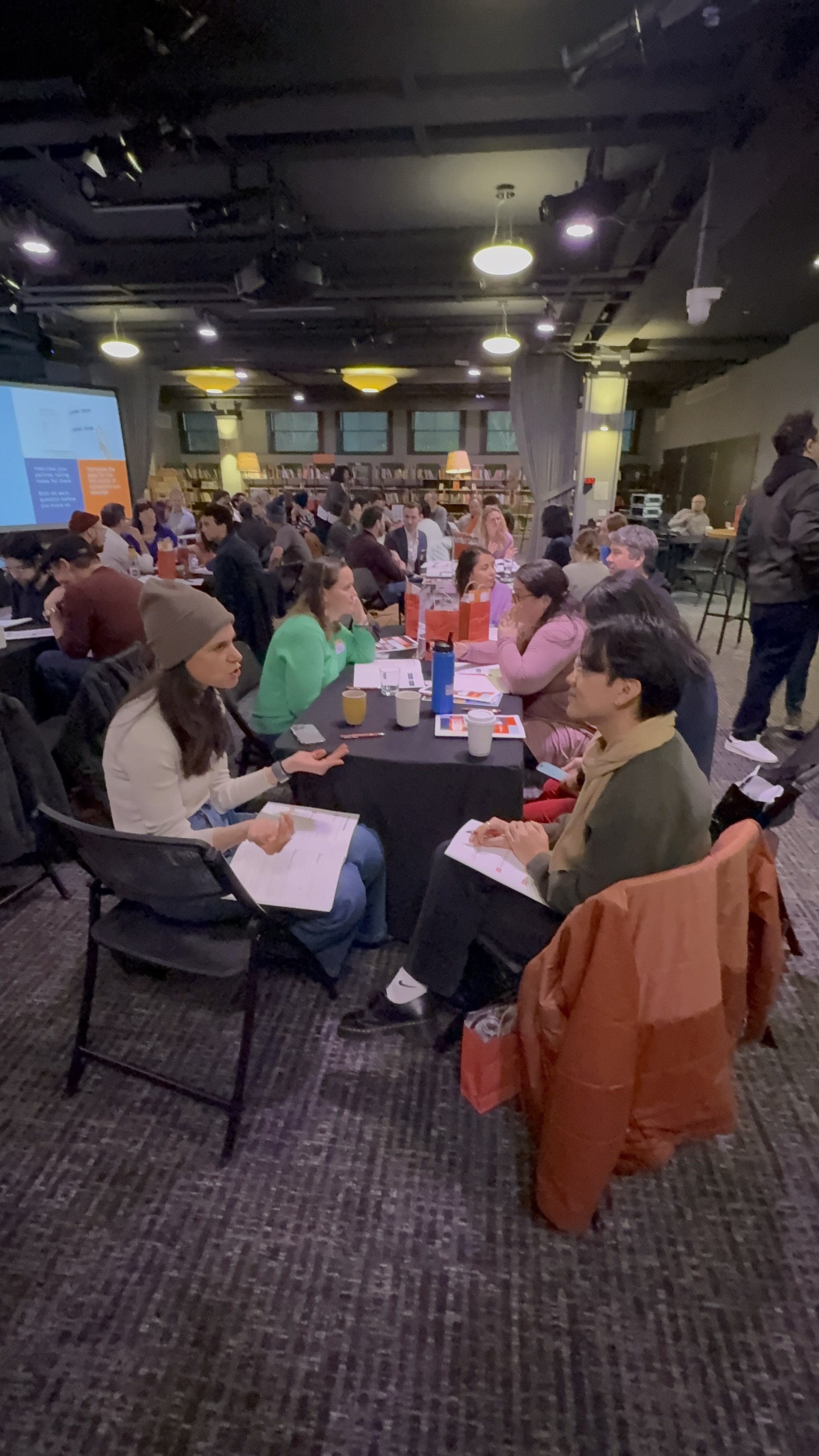 A large group of people seated around multiple tables in a dimly lit conference or workshop room with dark walls, bookshelves, and a large screen showing a presentation at the front. Attendees are engaged, taking notes, and having discussions.