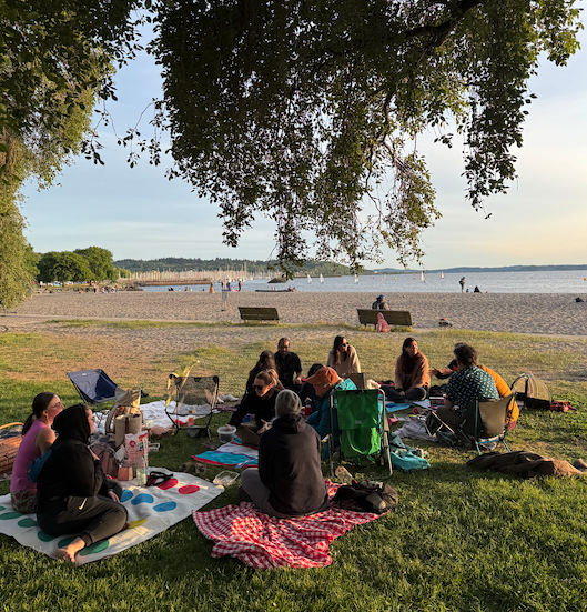 Group of people having a picnic under a tree by the beach with benches and sailboats in the background.