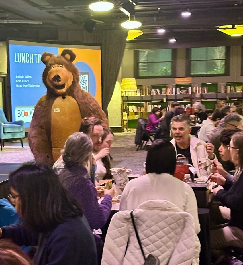 A person in a bear costume standing behind a group of people sitting at tables in a dining area. The background features bookshelves, windows, and a screen with a lunch promotion.