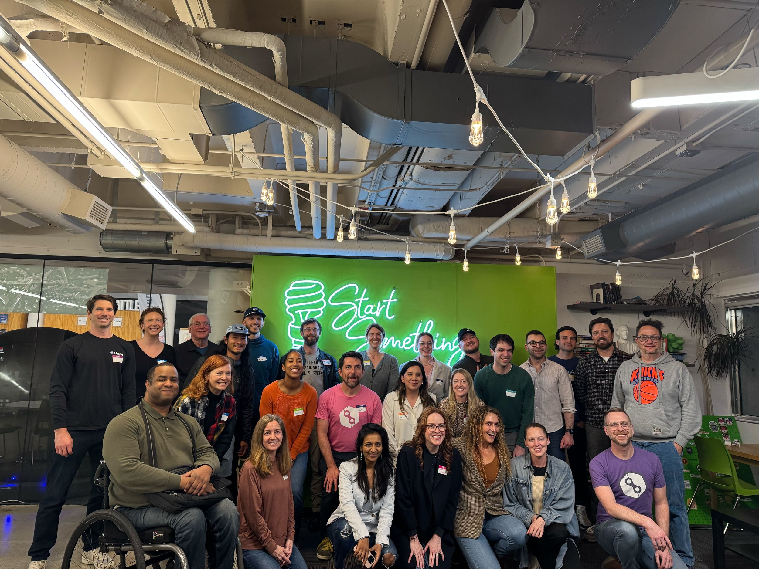A diverse group of people posing for a photo in an office space with a neon sign in the background that says 'Start Something' and a light fixture hanging from the ceiling.