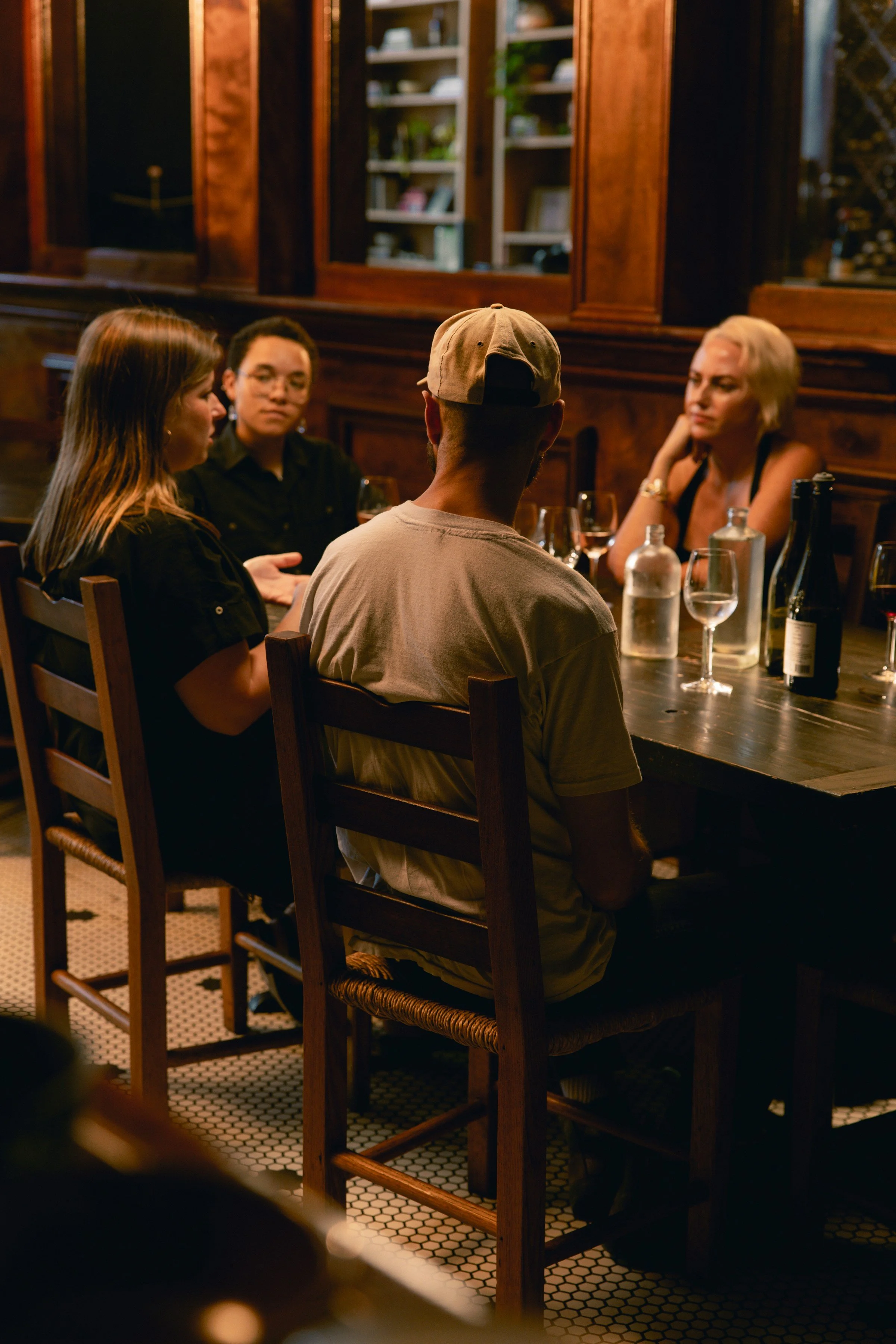 Four people sitting at a wooden table in a dimly lit restaurant, having a conversation with wine glasses and bottles on the table.
