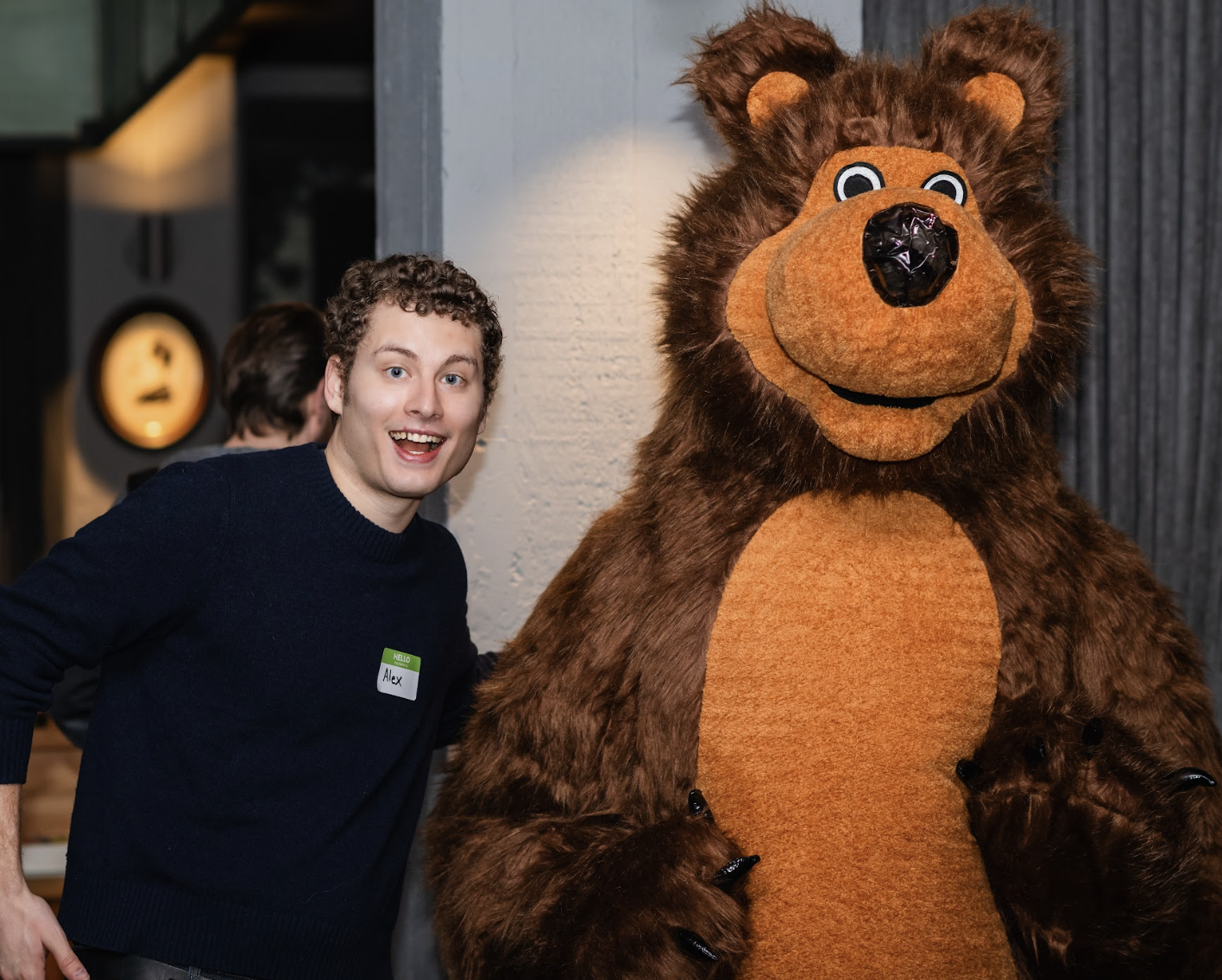 A young man with curly hair and a name tag that reads Alex standing next to a person in a bear costume.