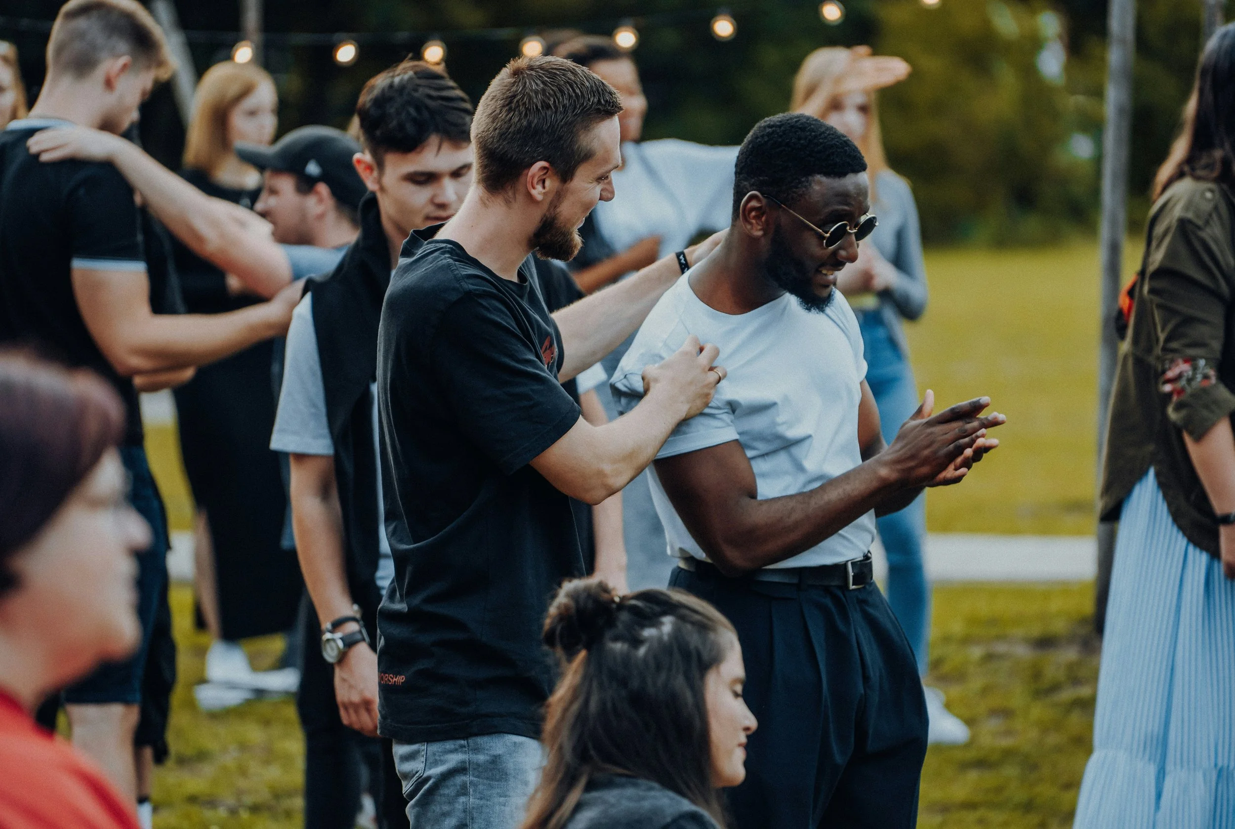 Group of people gathered outdoors, some are praying with hands clasped and heads bowed, others are touching shoulders or standing with eyes closed, during a spiritual or community event at sunset