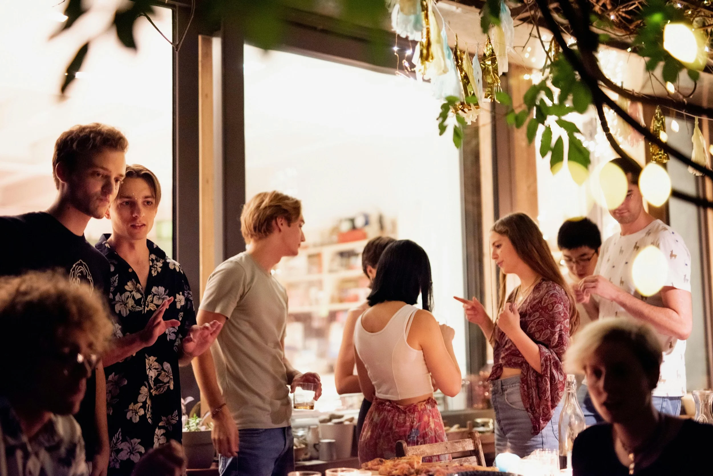 People socializing at a party or gathering indoors, with string lights and decorations hanging from the ceiling, some holding drinks and engaging in conversation.