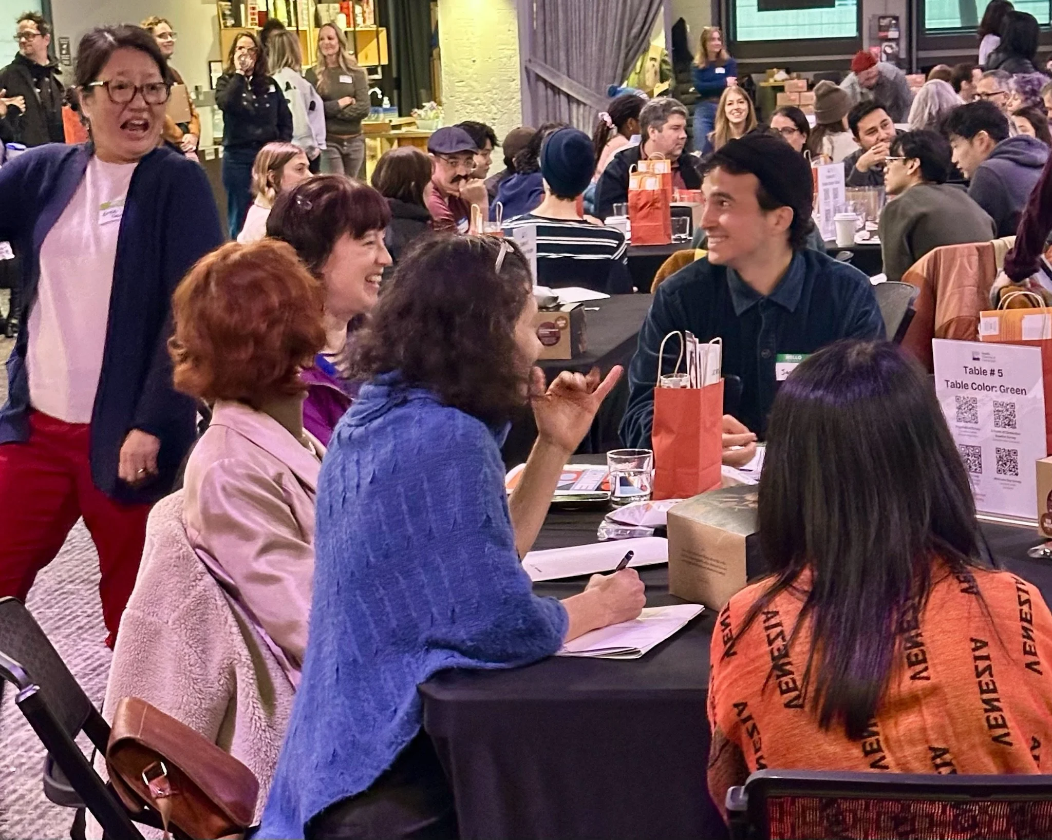 Group of people sitting at a round table engaged in conversation, with some standing behind them, in a lively indoor event setting.