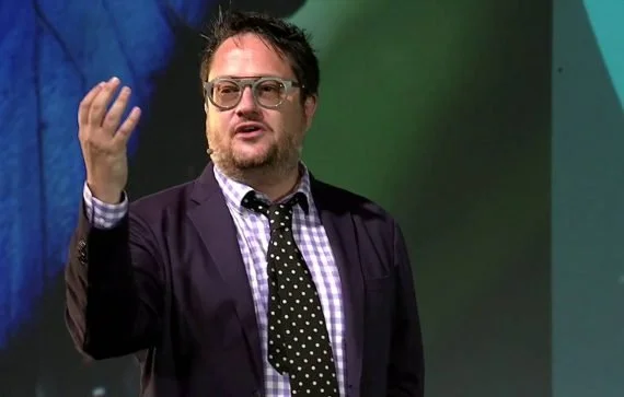 Man with glasses wearing a dark blazer, checkered shirt, and polka-dot tie, gesturing with his right hand while speaking.