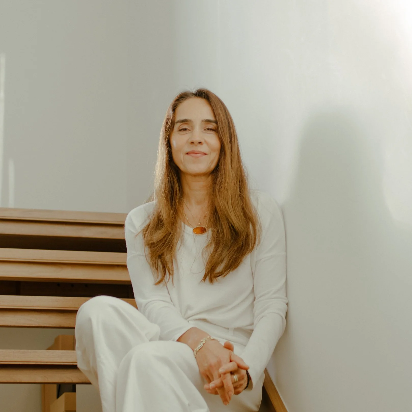 A woman with long brown hair, wearing a white outfit, sitting on stairs against a plain, light-colored wall.