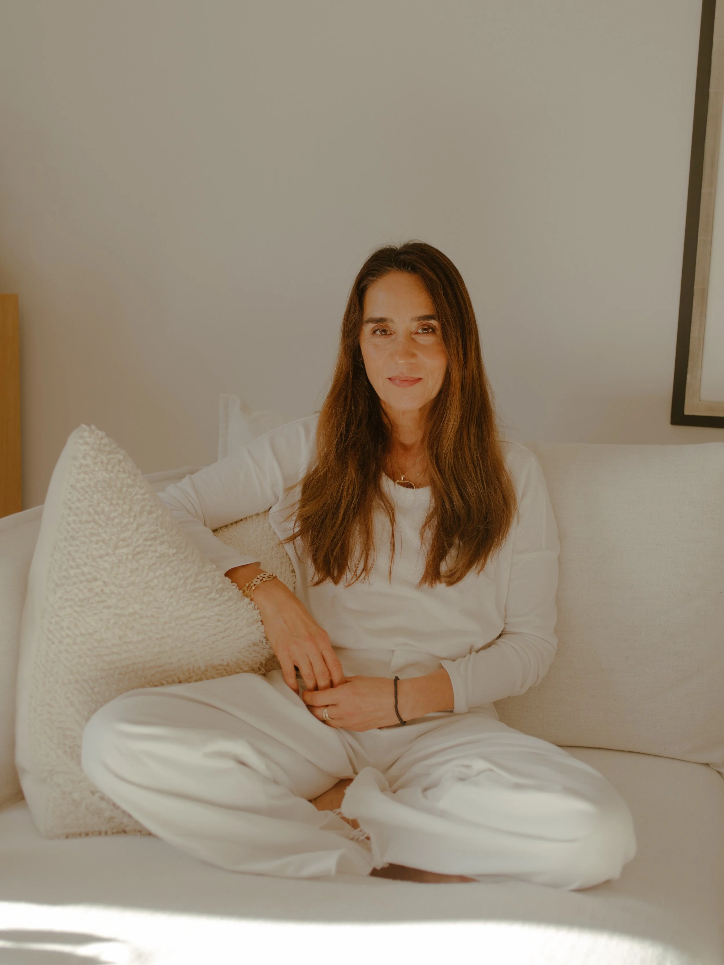 A woman with long brown hair, dressed in white, sitting cross-legged on a white couch, smiling softly at the camera, in a minimalistic indoor setting.