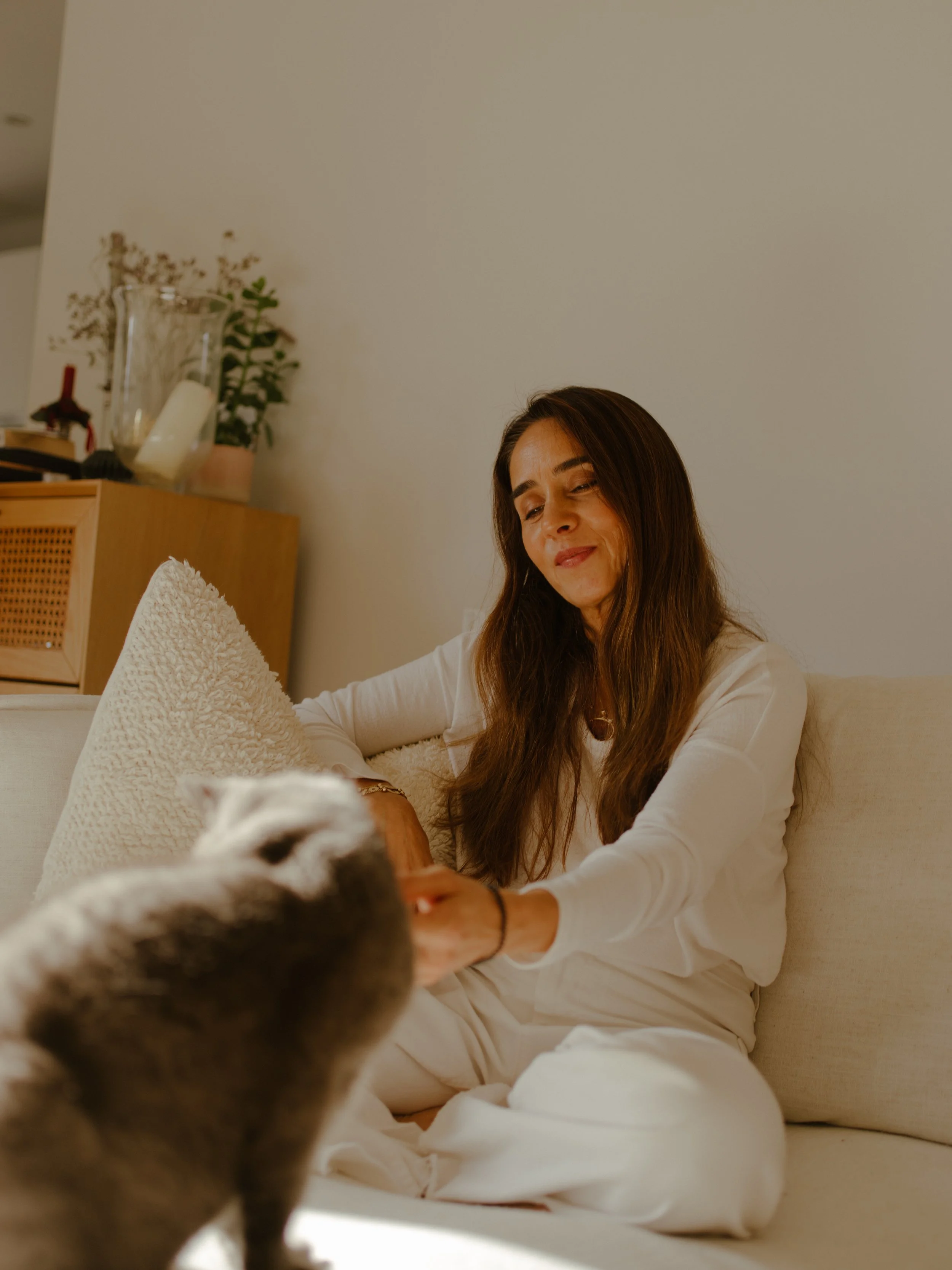 A woman with long brown hair wearing a white outfit sitting on a beige couch, smiling while interacting with a black and white cat, in a cozy living room.