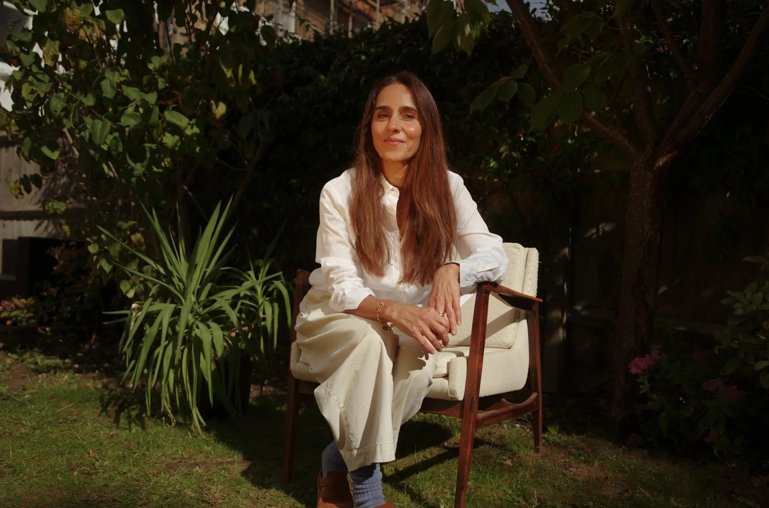 A woman with long brown hair sitting outdoors in a garden on a wooden and cream-colored armchair, wearing a white shirt and cream pants, with plants and trees in the background.