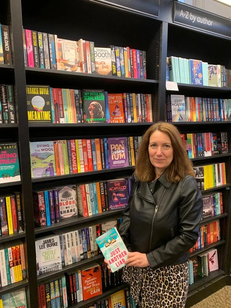 A woman standing in front of a bookshelf filled with books at a bookstore or library, holding a book titled 'Just Another Liar'. The bookshelf has a sign labeled 'A-Z by author' at the top.