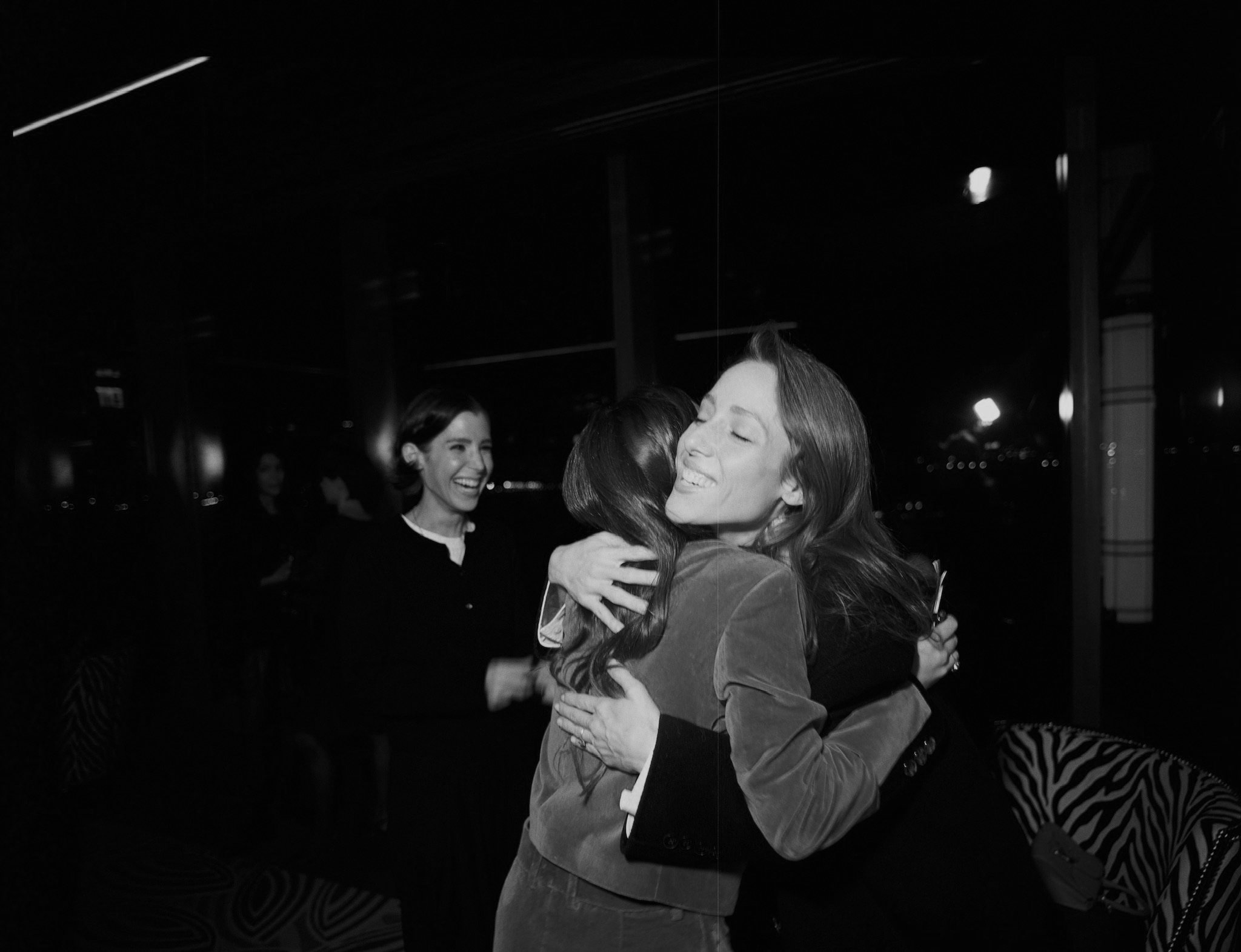 Two women hug each other in a black-and-white photo, with a third woman smiling in the background at a social gathering.