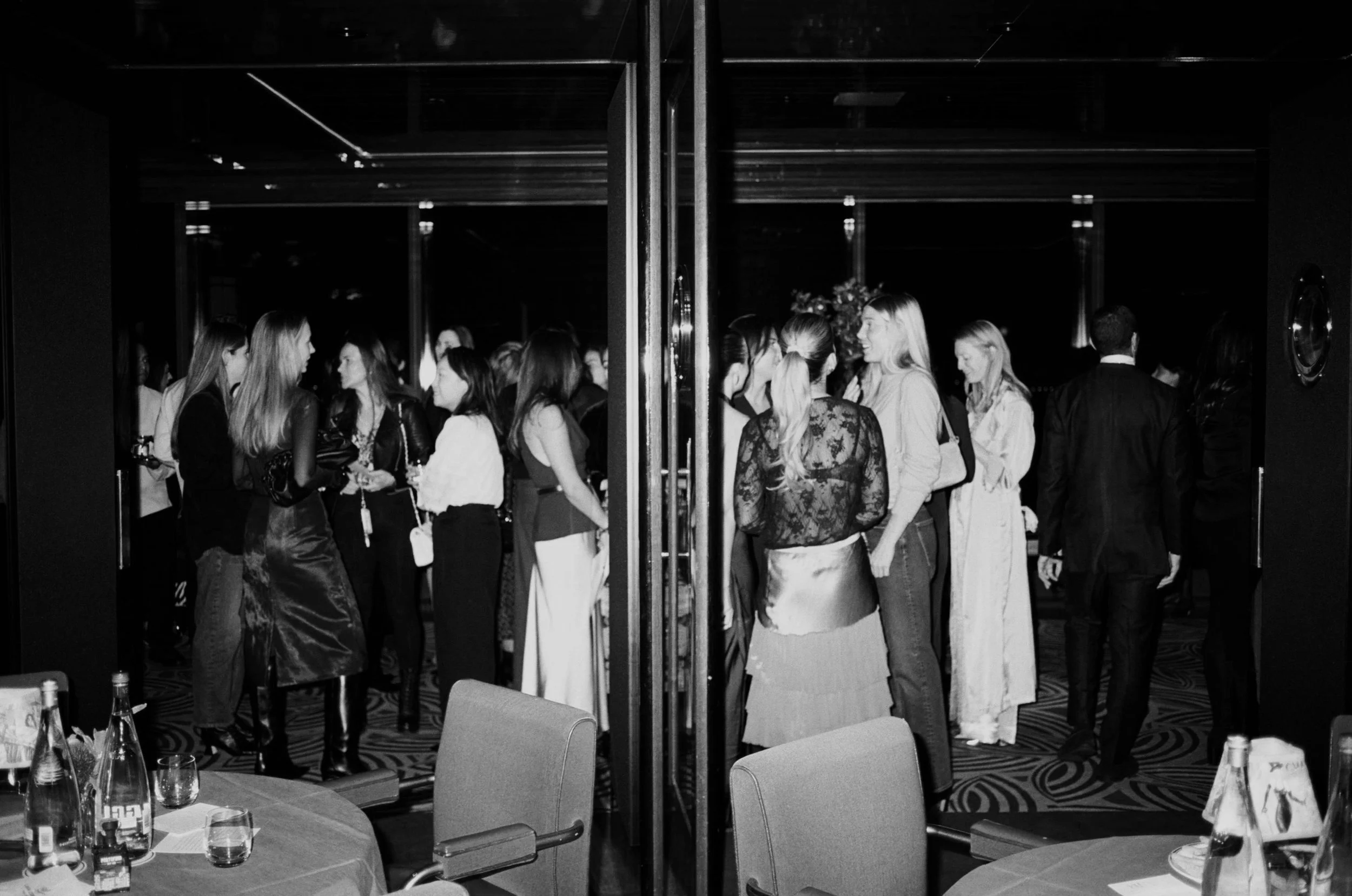 People gather in a dimly lit room, socializing at an event, separated by a glass divider.