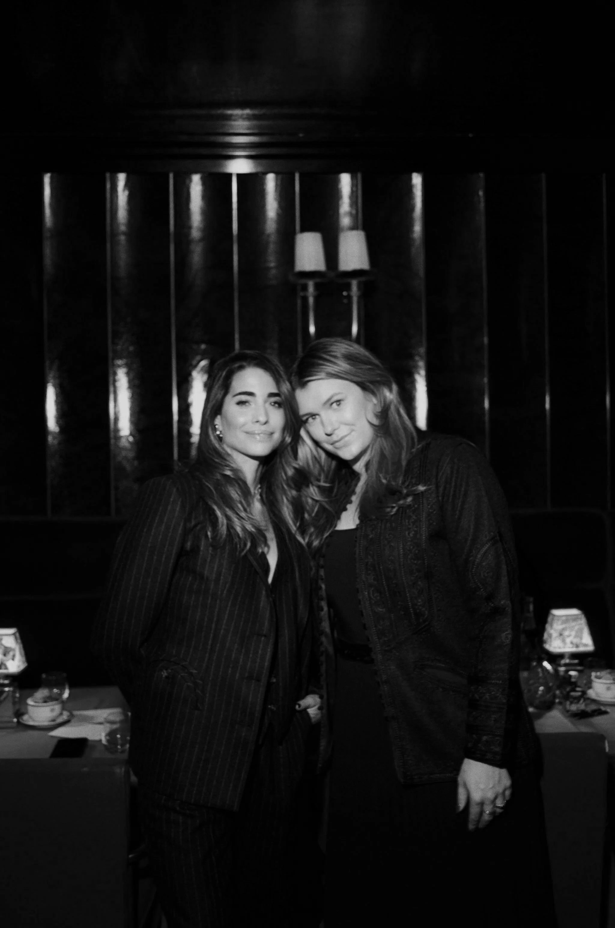 Two women standing close together indoors, smiling, with a dark, reflective wall and a table with cups and bottles in the background.
