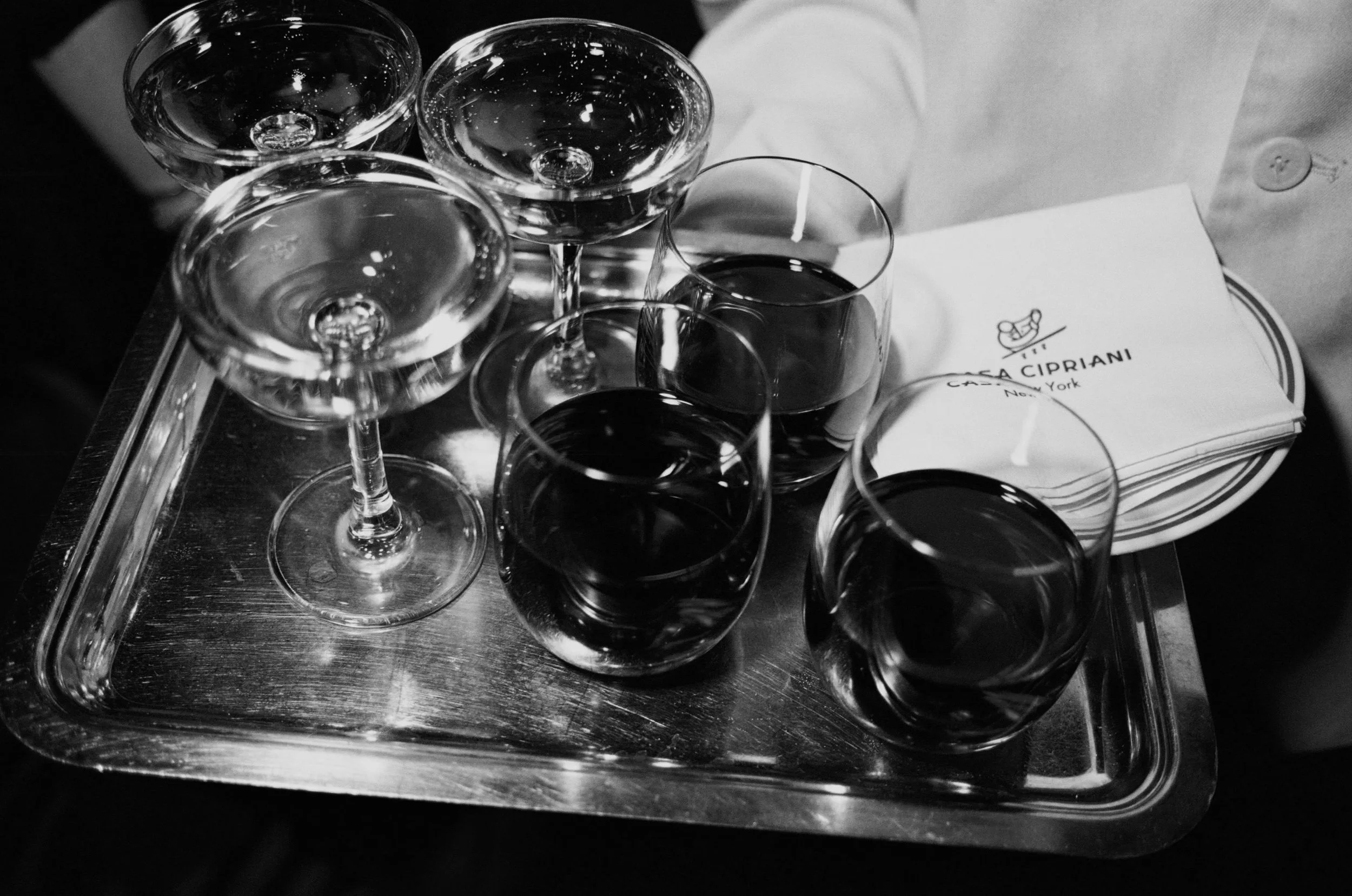 A tray holding multiple wine glasses with a dark liquid, likely red wine, along with several empty glasses and a napkin with the logo and name "Casa Cipriani" from New York.