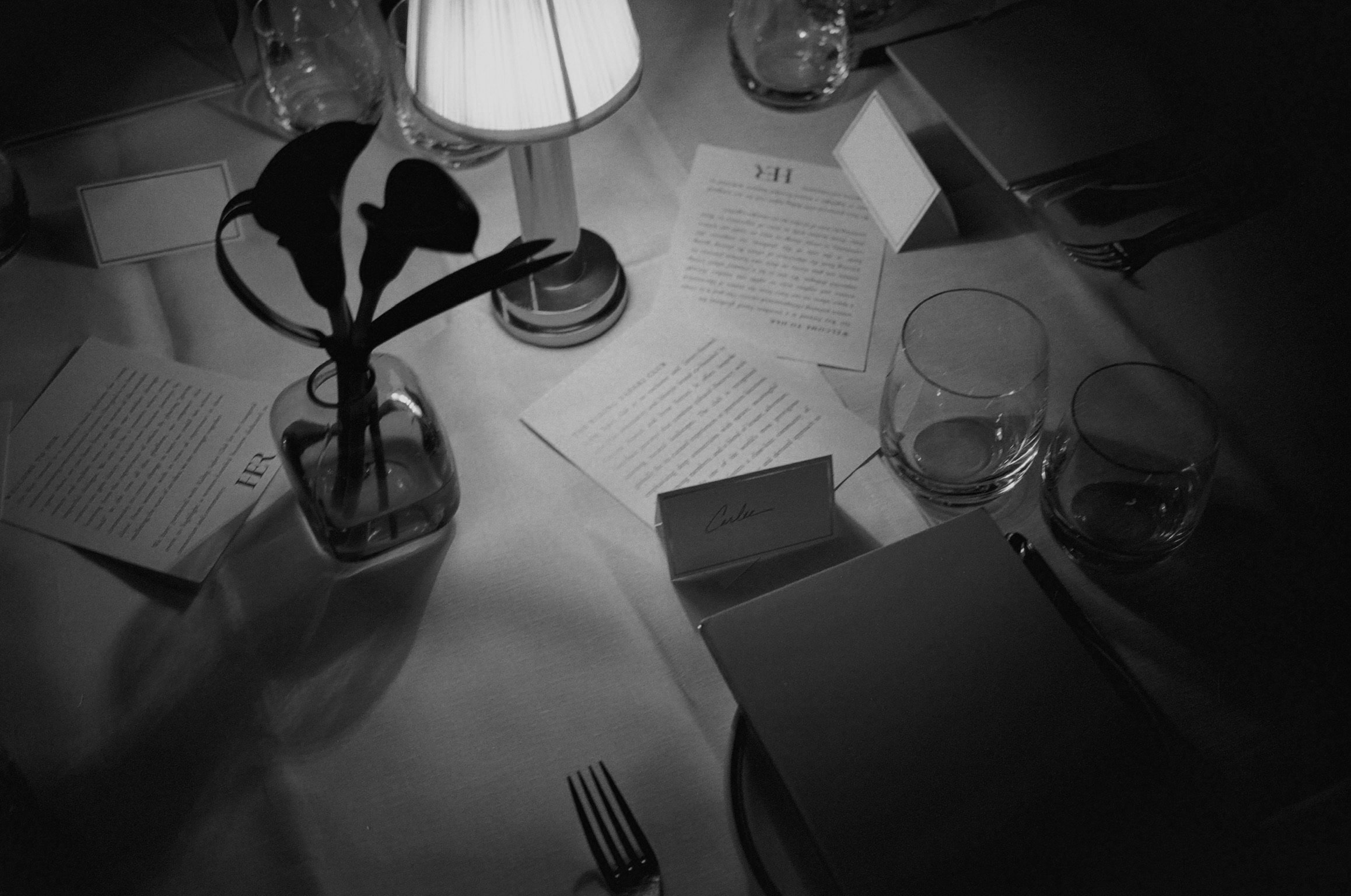 A black and white photograph of a table setting at a formal event. The table has a small vase with tulips, a candle, glasses, and various written notes and cards. The setting suggests an elegant or intimate gathering.