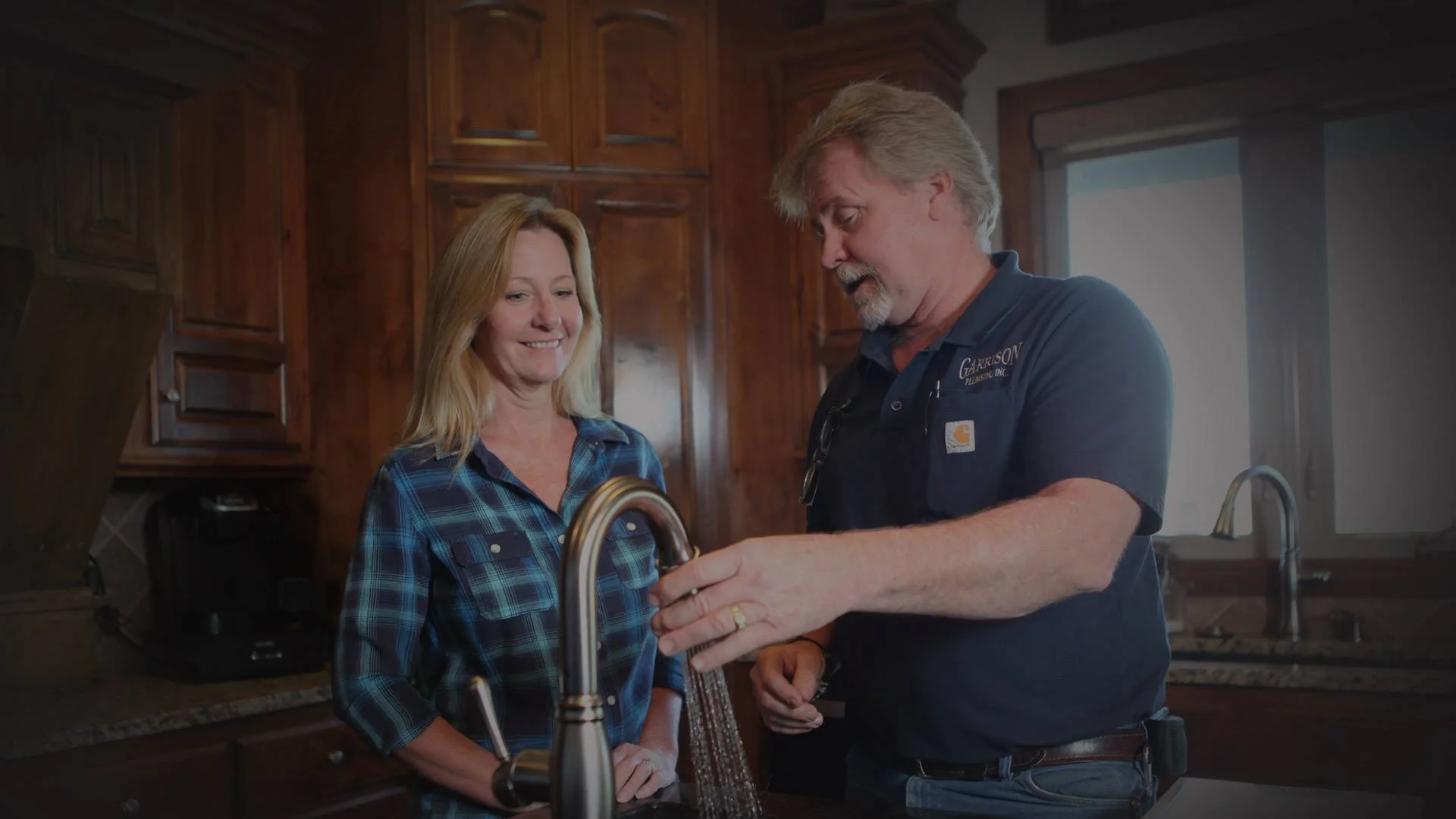 A man and woman in a kitchen, with the man showing the woman a fixture at the sink.