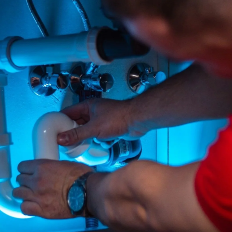 A person inspecting or working on plumbing pipes under a sink, with blue lighting.