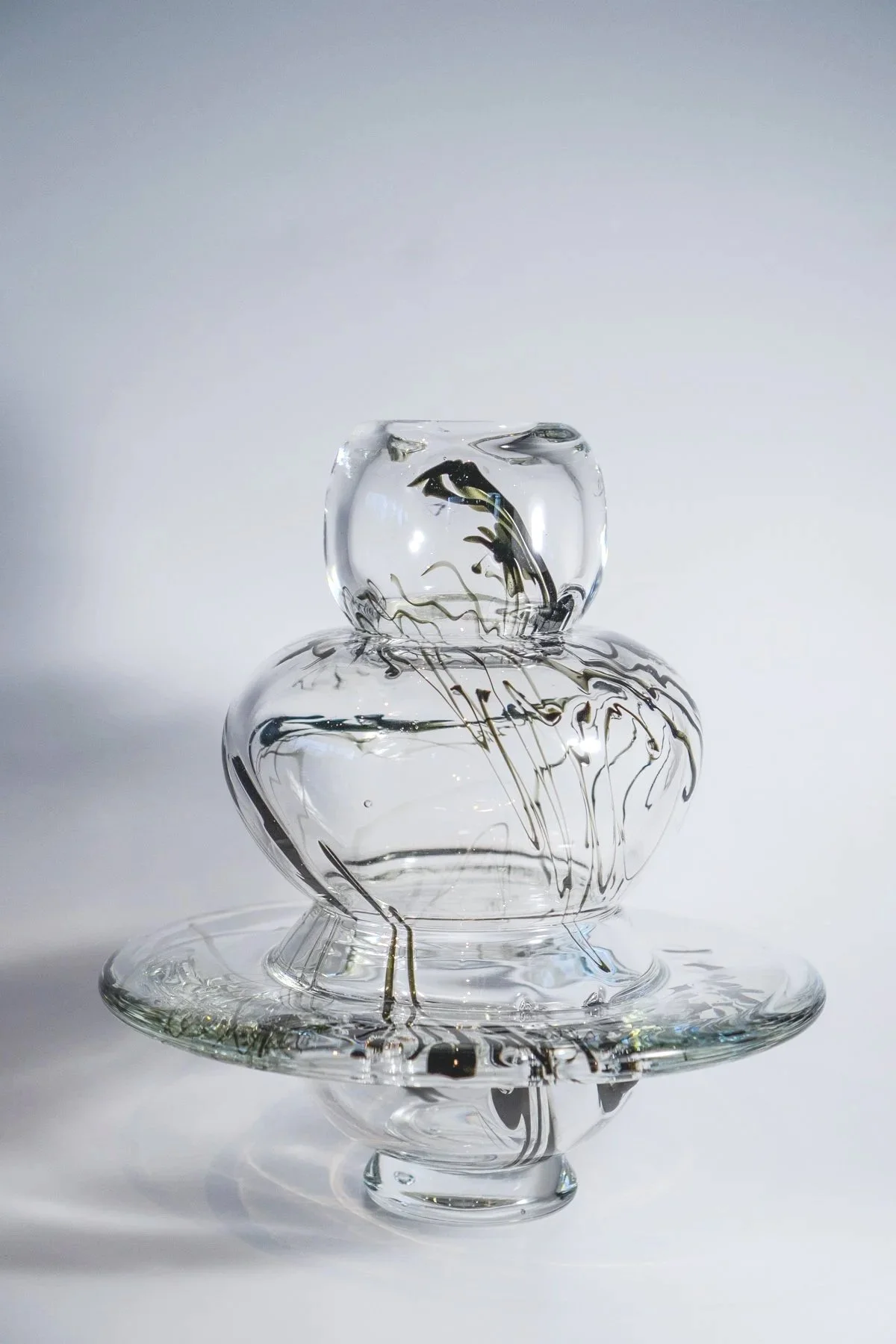 A glass sculpture resembling a snowman, with a rounded top and a larger rounded base, decorated with black streaks and sitting on a wide glass plate, against a plain gray background.