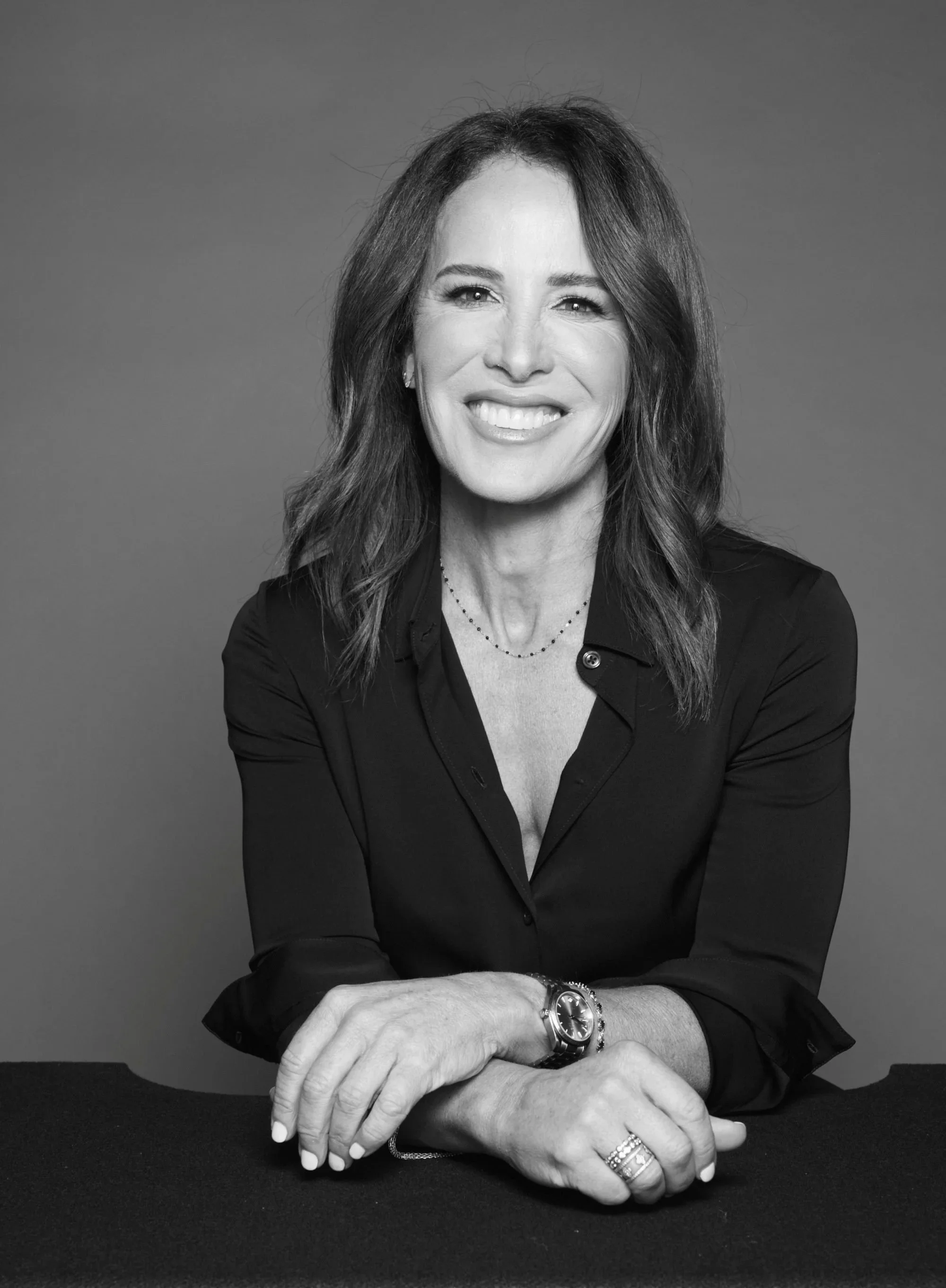 Black and white portrait of a woman, Dr. Sarah de la Torre, with shoulder-length hair, smiling, wearing a dark shirt, a watch, rings, and a necklace, sitting at a table with crossed hands.