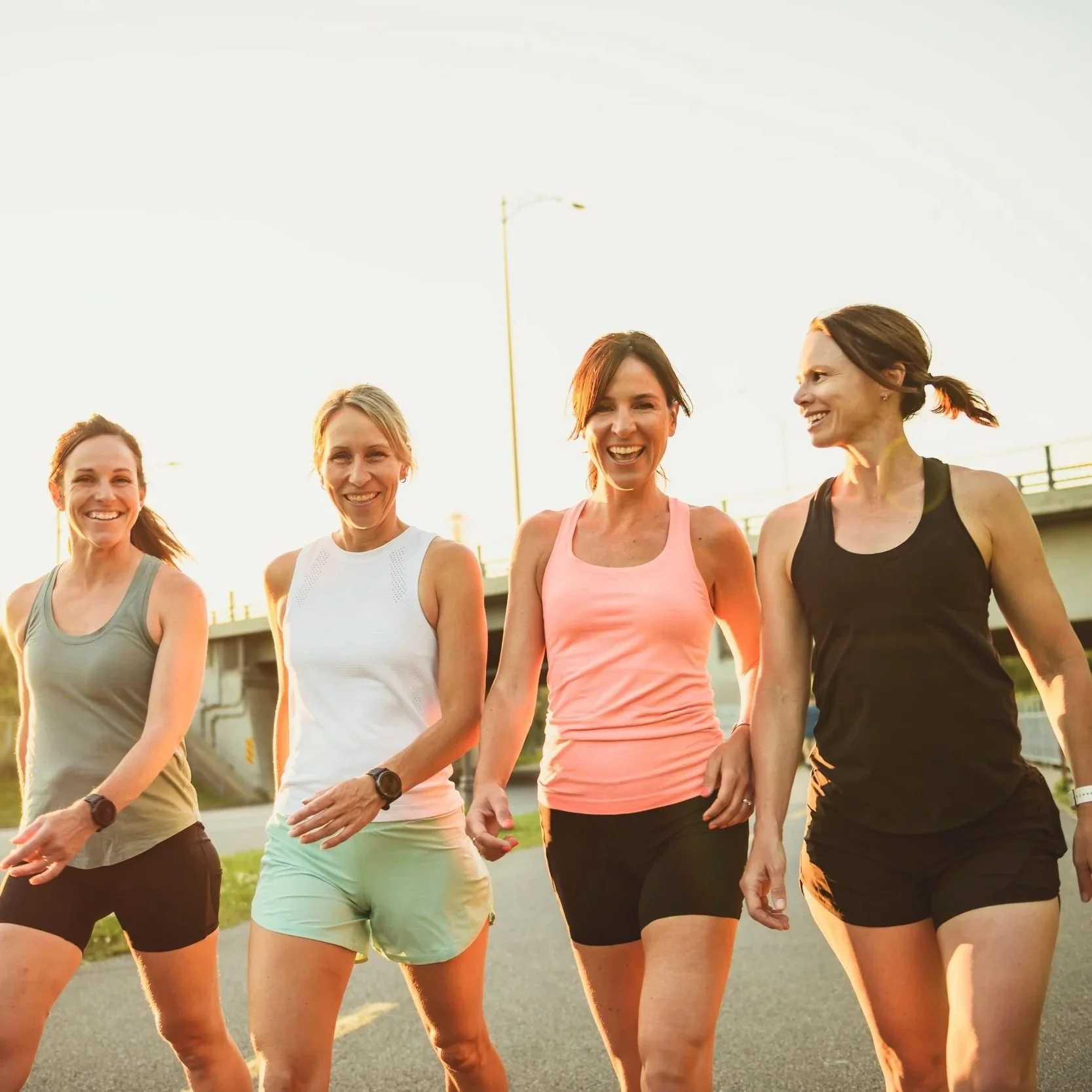 Four women in their 40s in athletic clothing walking outdoors during sunset, smiling and chatting.