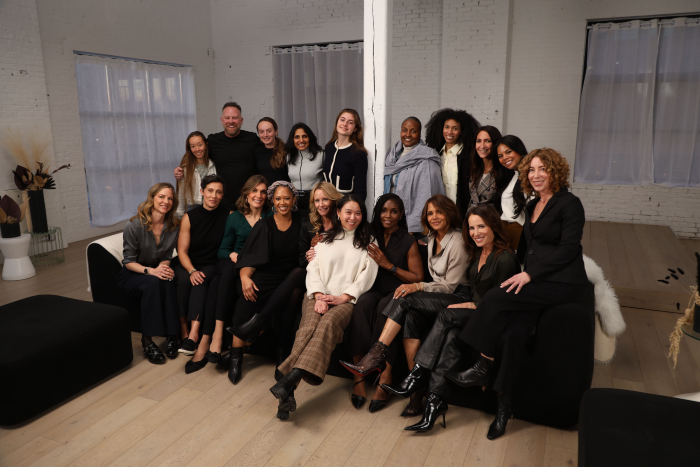 Group of 17 people, mostly midlife women, posing together in a bright room with white brick walls and large windows.