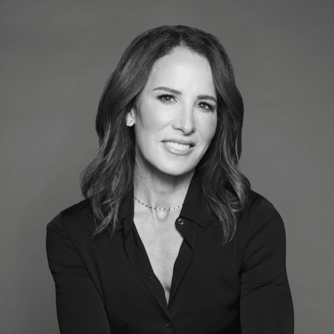 Black and white portrait of a woman, Dr. Sarah de la Torre, with shoulder-length wavy hair, smiling, wearing a dark blouse and a delicate necklace, against a plain background.