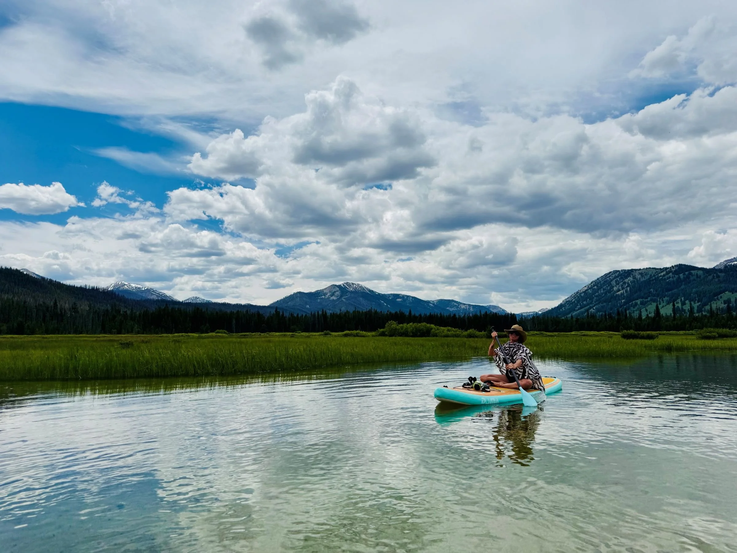 A midlife woman sitting on a paddleboard paddling in a river in Idaho with mountains and cloudy sky in the background.