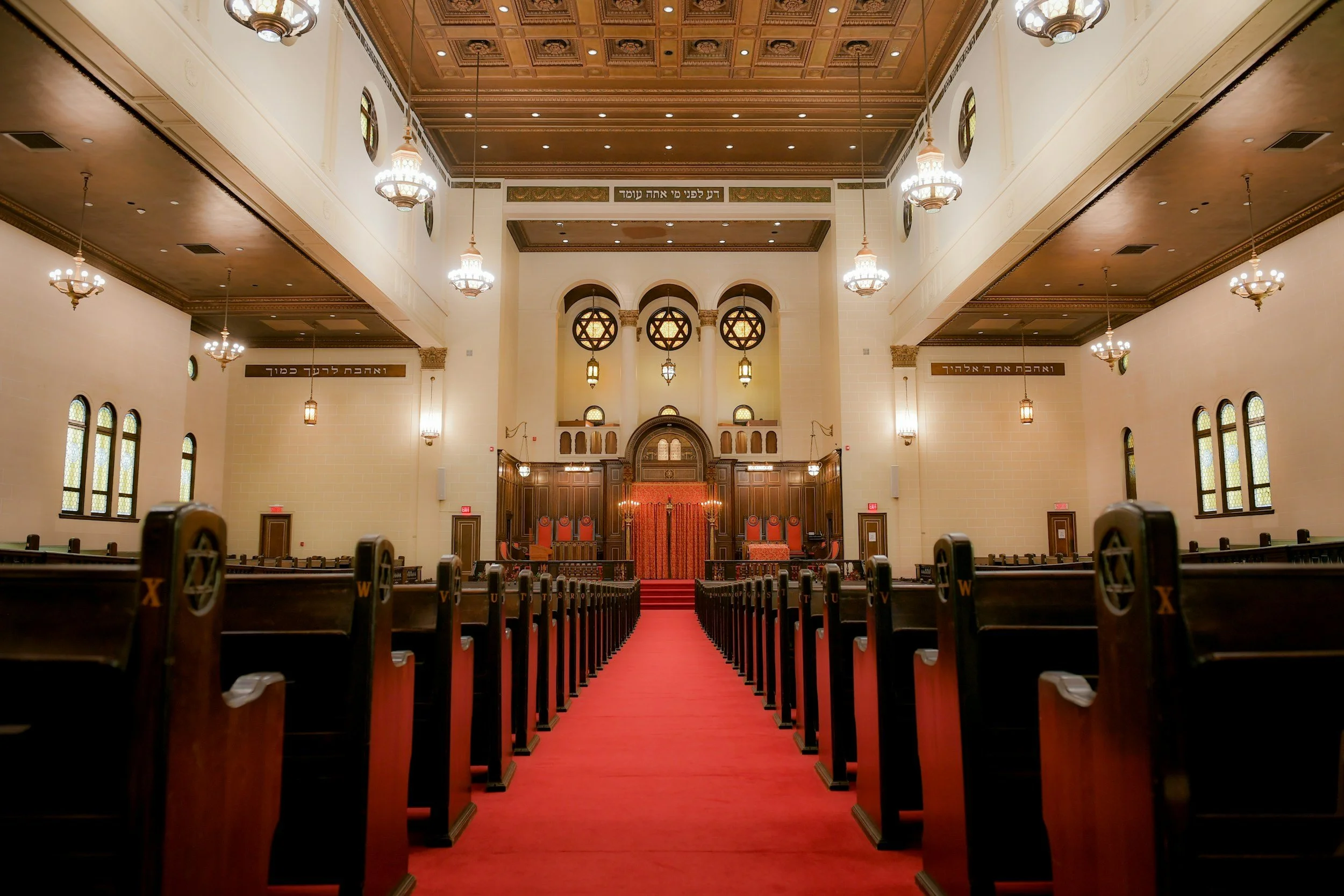 Interior of a synagogue with wooden pews, a red carpet, and an elevated ark at the front, decorated with Hebrew symbols and inscriptions.