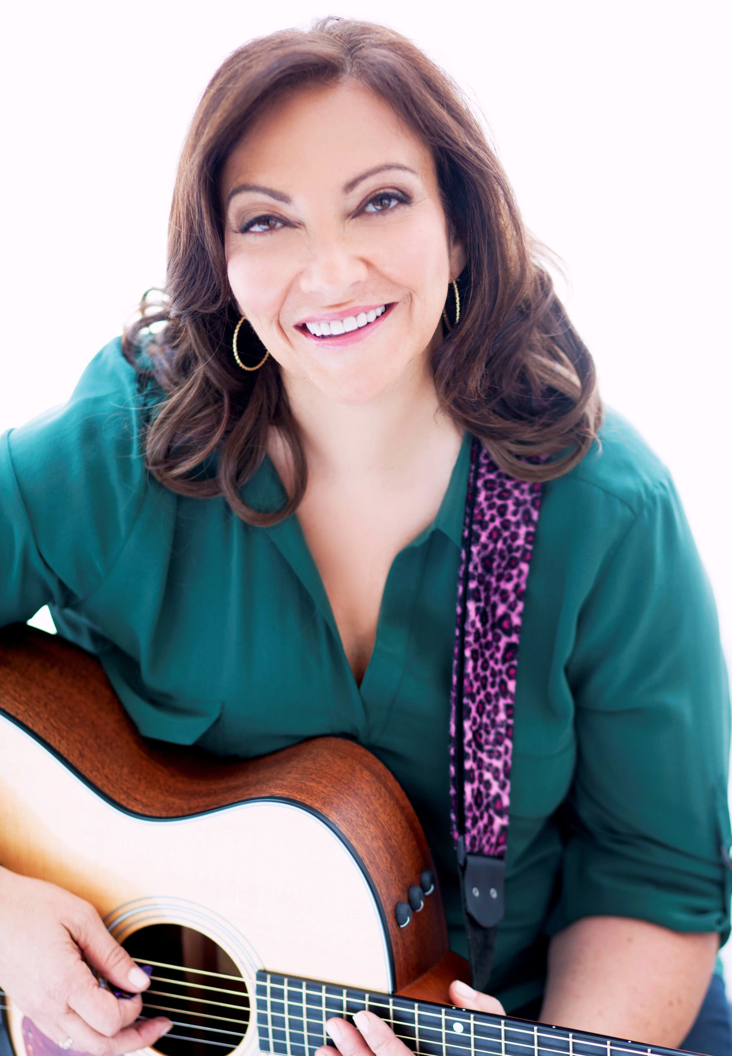 A woman with brown hair, smiling, holding an acoustic guitar, wearing a teal shirt with purple leopard print accents and hoop earrings.