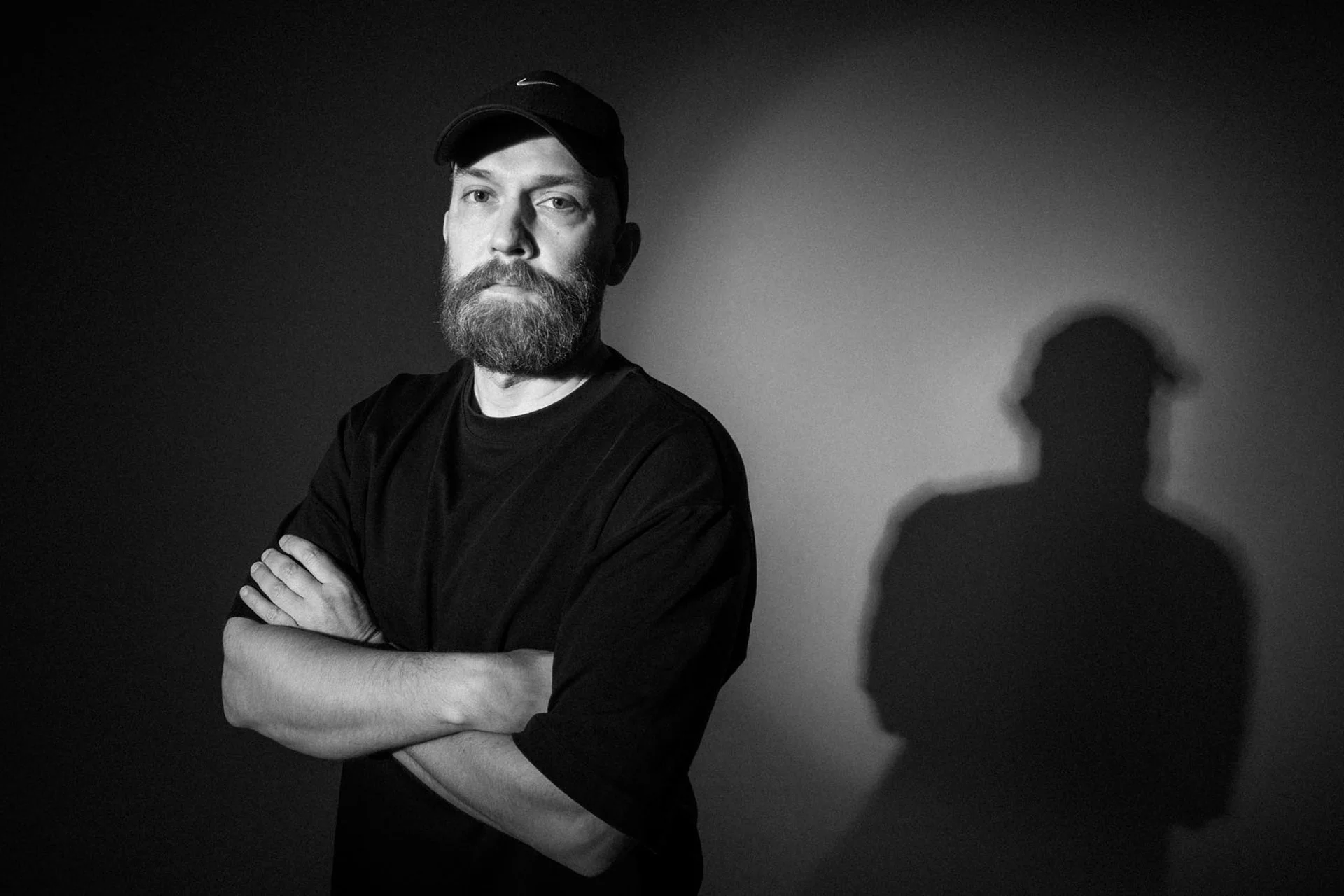 Schwarzweißporträt eines Mannes mit Bart und Cap, der die Arme verschränkt vor einer dunklen Wand steht. Sein Schatten ist deutlich an der Wand sichtbar.