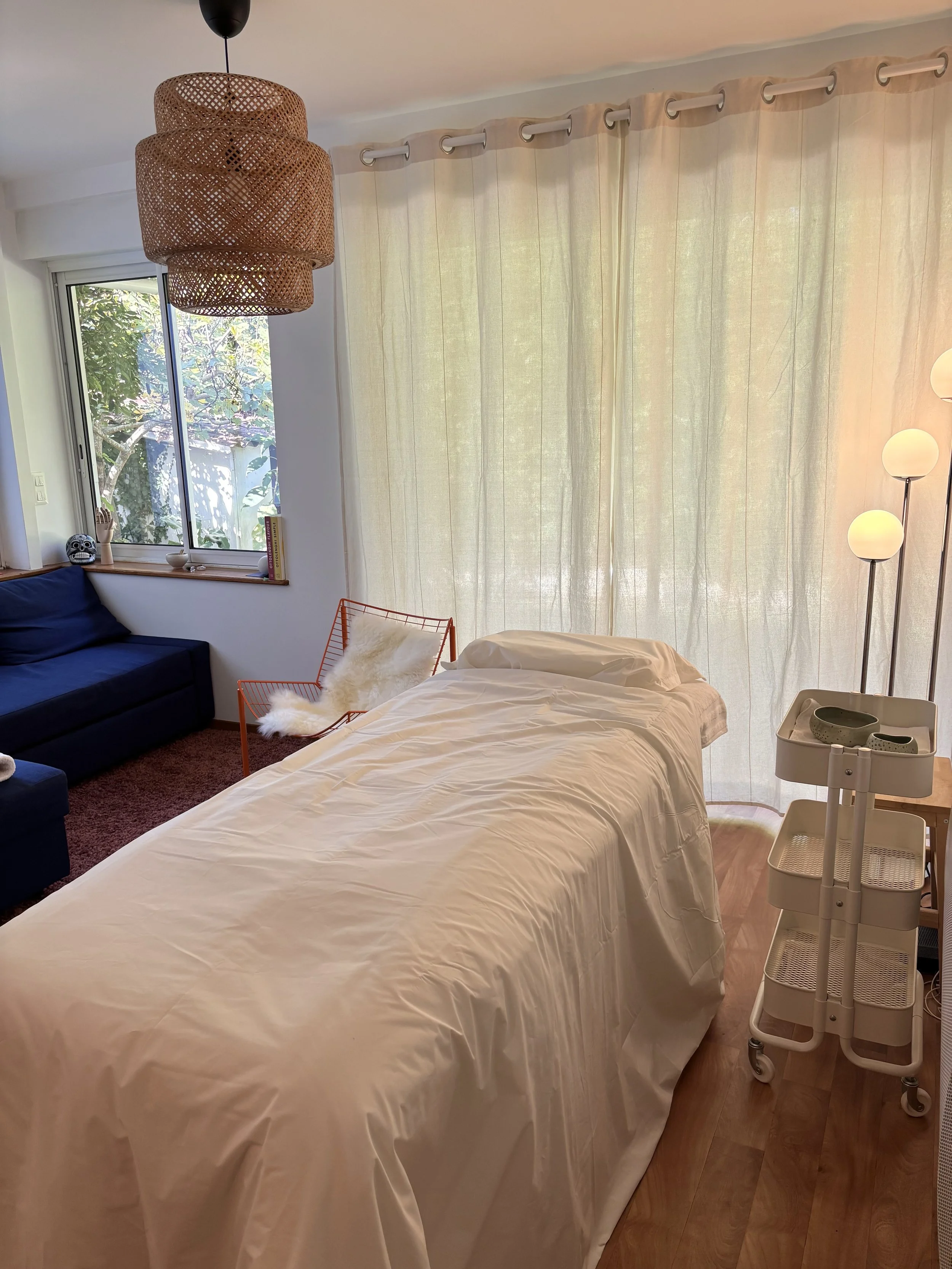 Une pièce lumineuse avec un lit, des rideaux en voile, un fauteuil orange avec un coussin en fourrure blanche, un canapé bleu, une étagère avec un crâne décoratif et des livres, une lampe sur pied avec plusieurs globes, et une lumière suspendue en rotin