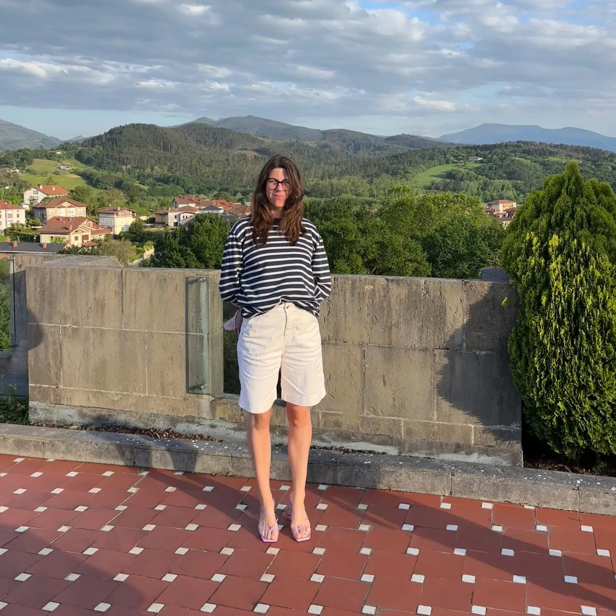 Une femme en vêtements décontractés, portant un t-shirt à rayures noires et blanches, et un short beige, se tient sur une terrasse avec un paysage de montagnes, de forêts et de maisons en arrière-plan.
