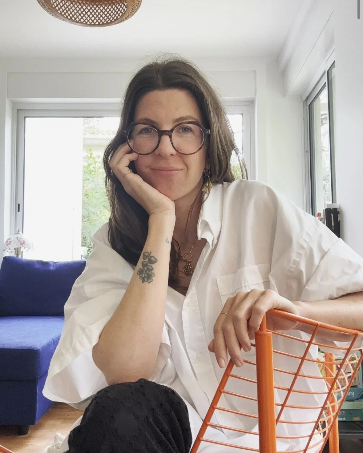 Une femme aux cheveux bruns, portant des lunettes, assise à une table dans un intérieur lumineux avec de grandes fenêtres. Elle porte une chemise blanche et regarde l'objectif avec un léger sourire. Elle a un tatouage sur le bras gauche et tient un panier orange en métal.