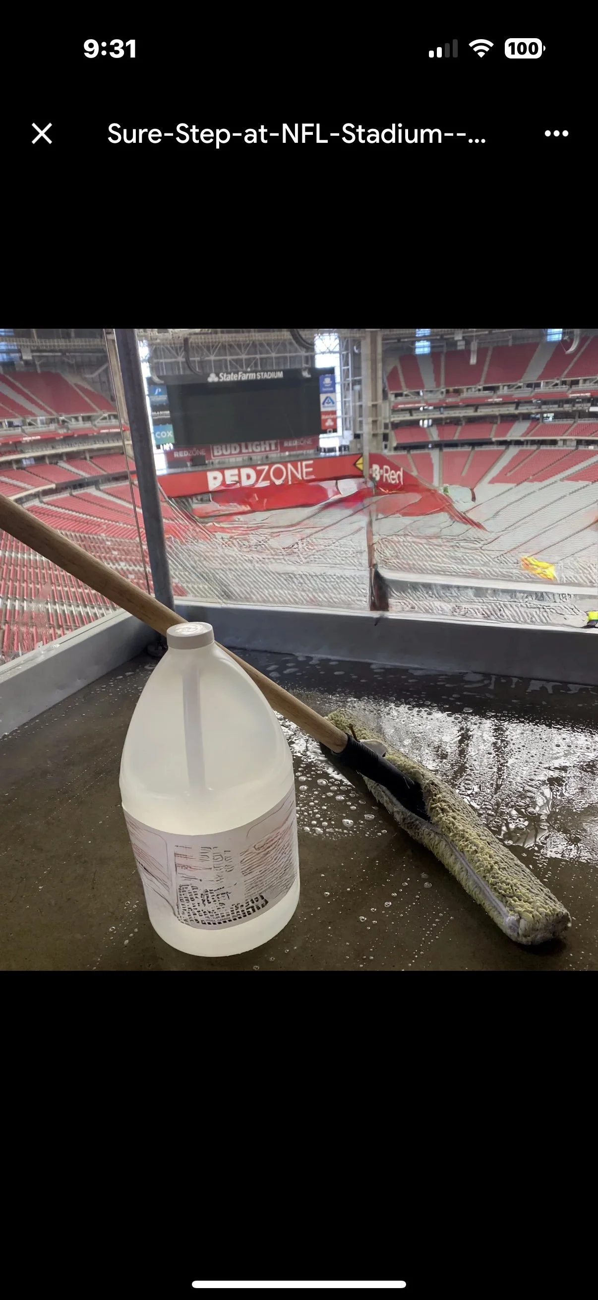 Cleaning supplies, including a large plastic jug and a scrub brush, on a window ledge overlooking an empty football stadium with red seats and a large scoreboard after slip patrol non slip treatment.