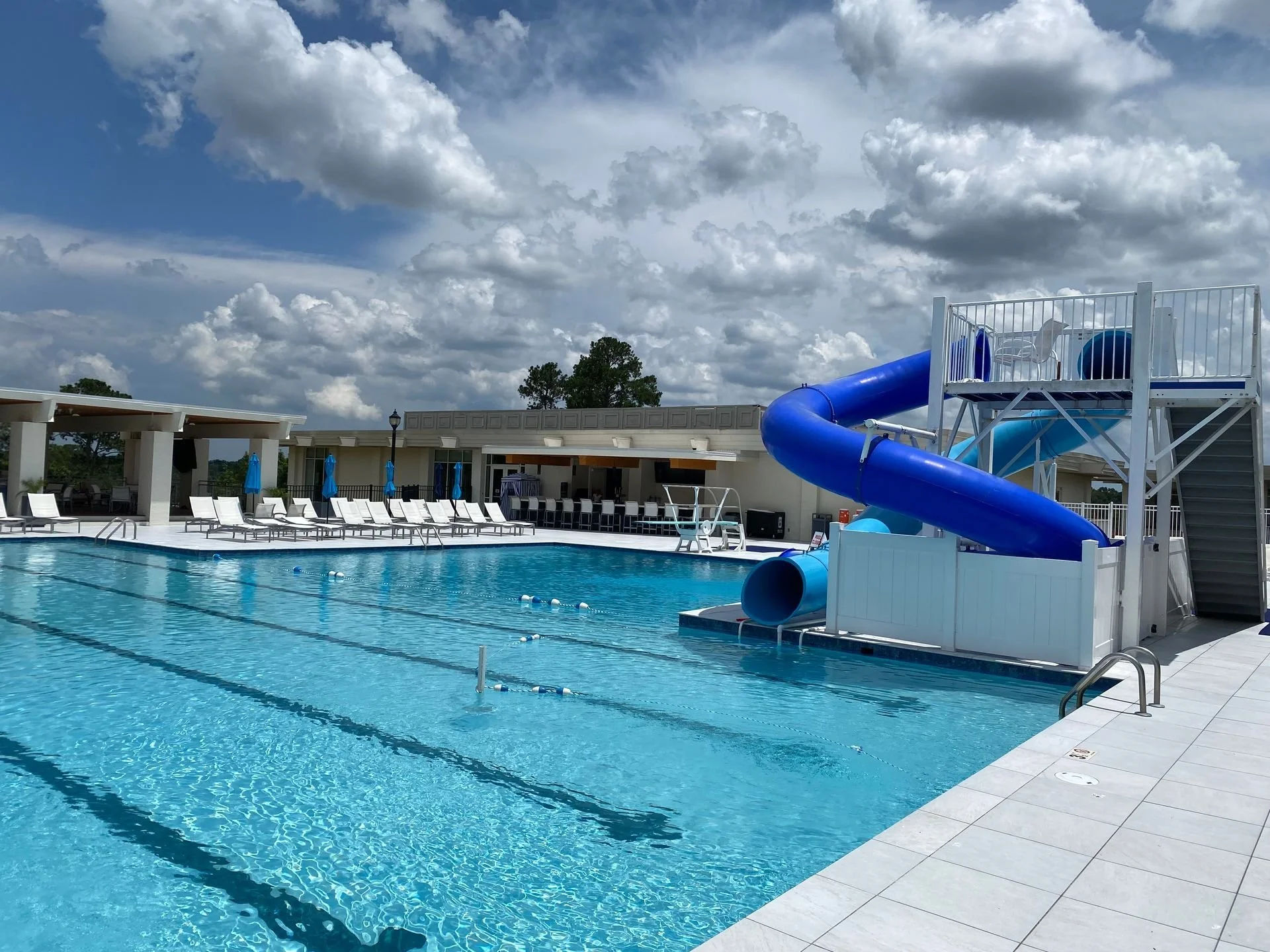 Empty outdoor swimming pool with blue waters, a water slide, and lounge chairs under a cloudy sky after slip patrol non slip treatment. 