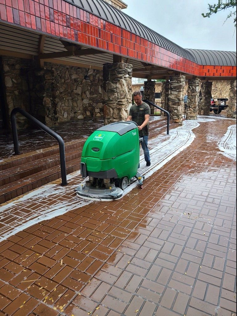A man operating a green ride-on floor scrubber cleaning a wet brick sidewalk outdoors after slip patrol non slip treatment. 
