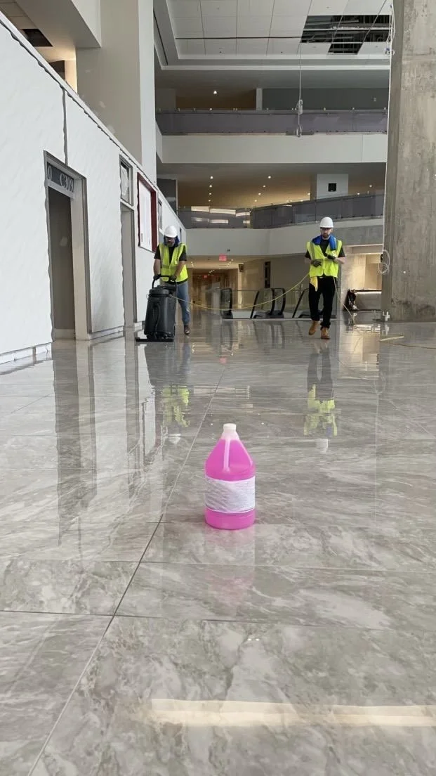 Workers in safety vests and helmets cleaning the shiny marble floor of a large building, with a pink and white bottle of cleaning solution in the foreground.