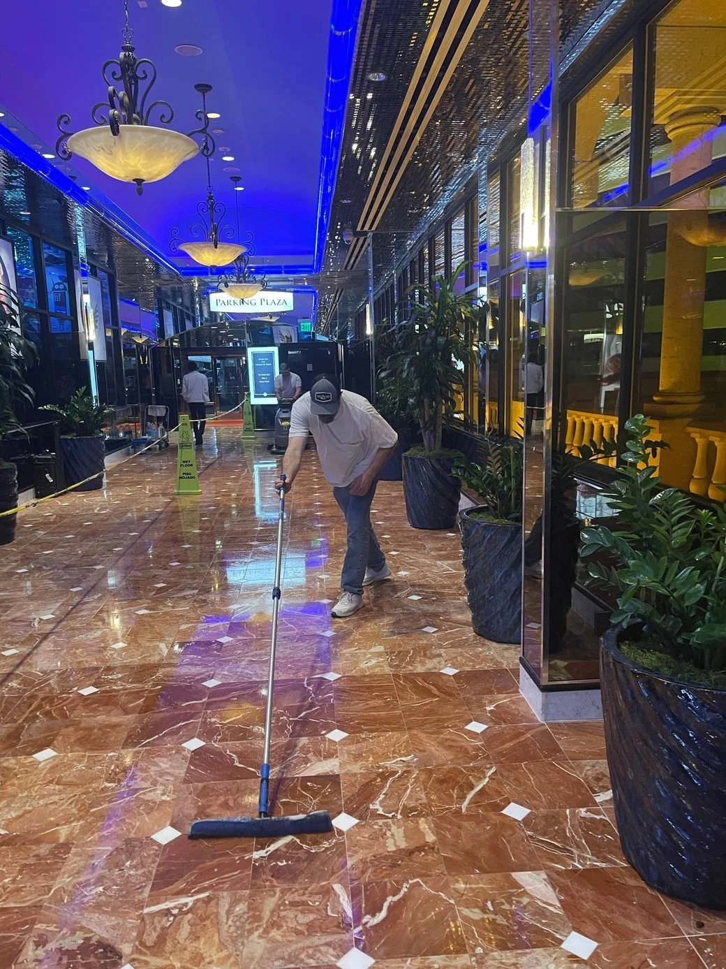A man cleaning the shiny brown marble floor of a hotel lobby or casino with a mop. There are large potted plants along the side and blue neon lights on the ceiling, with chandeliers hanging down. Several other people are in the background, and a sign reads 'Parking Plaza'.