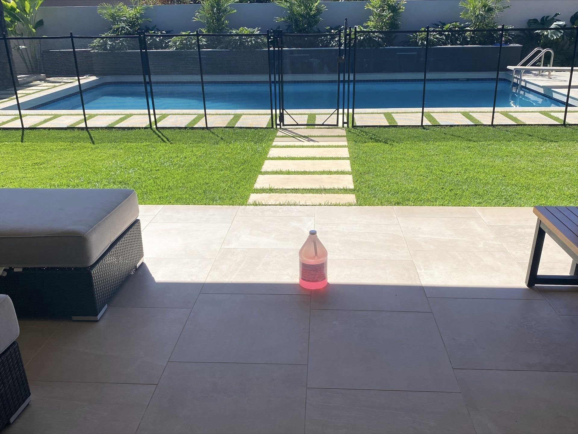 View of a backyard with a swimming pool surrounded by a safety fence, a grassy area with stepping stones leading to the pool, and patio furniture including a cushioned chair and a small table, with a family-sized bottle of pink-colored liquid on the patio.