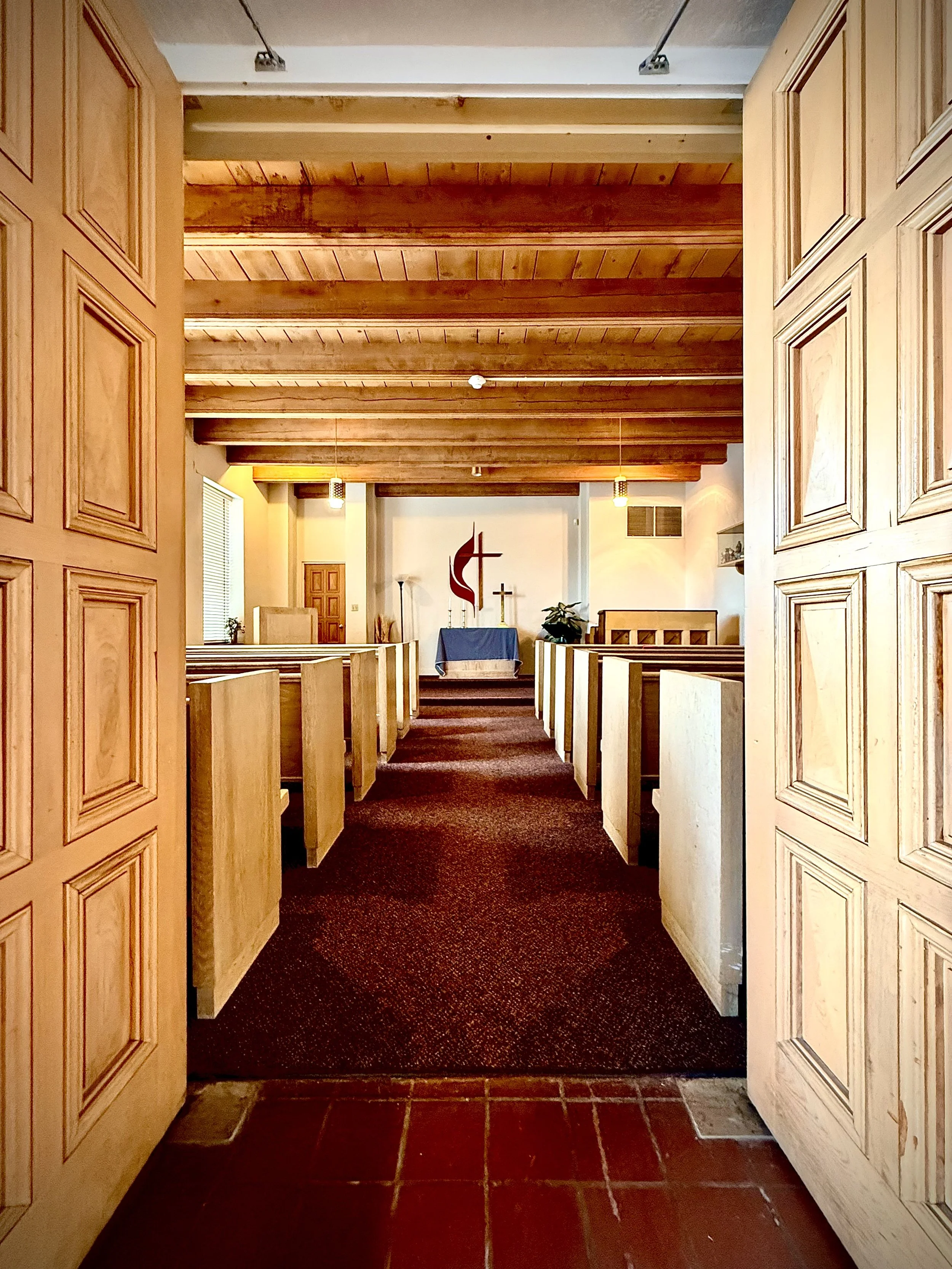 View through wooden double doors into the chapel interior with rows of pews, a carpeted aisle, altar, and religious symbols on the wall.