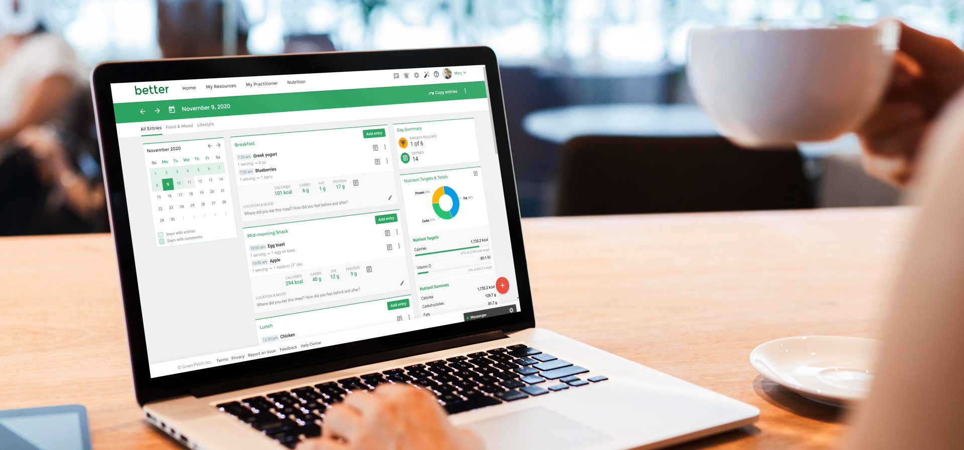 A person holding a white coffee cup in front of a laptop on a wooden table in a cafe.