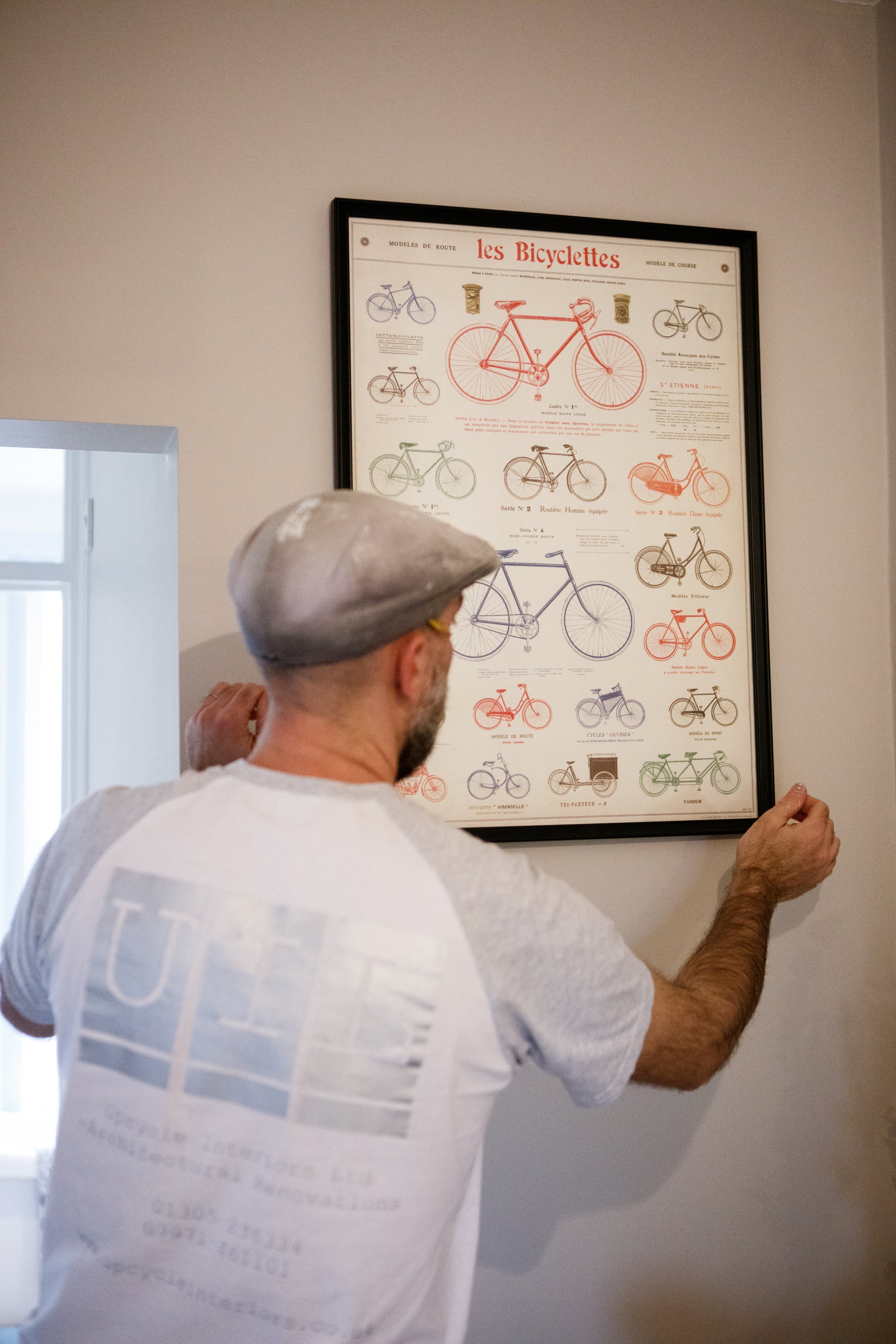 A man adjusts a framed chart of bicycle models on a wall.