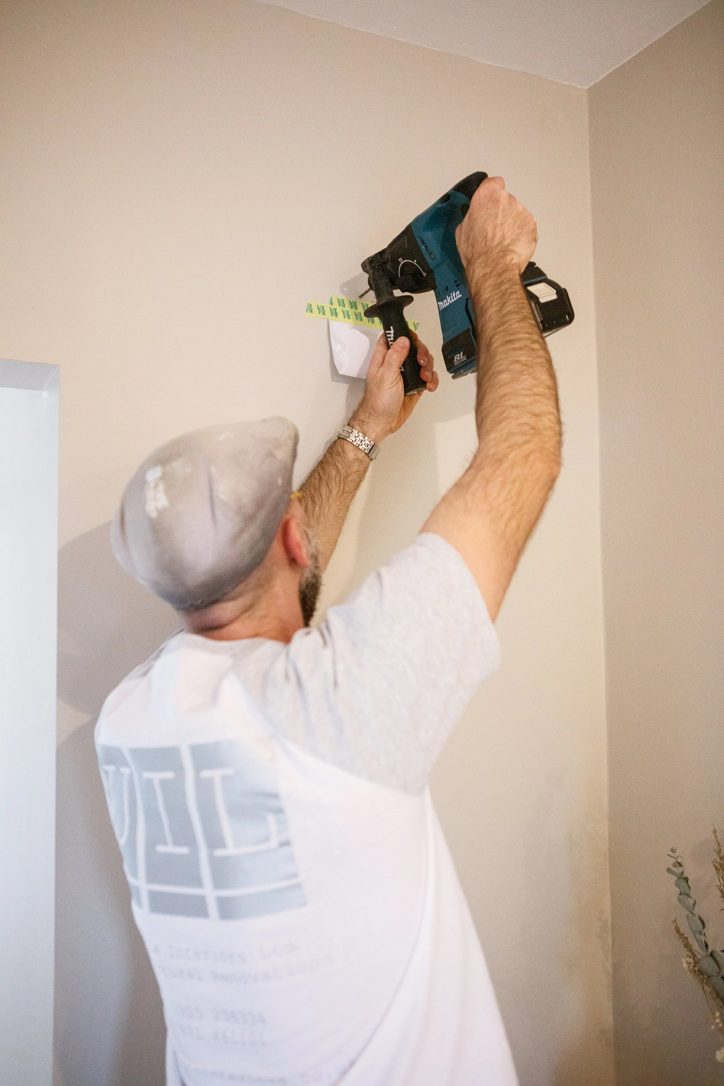 A man installing an item on a beige wall, using a cordless power drill. He is wearing a gray cap and a white t-shirt, and is focused on the task.