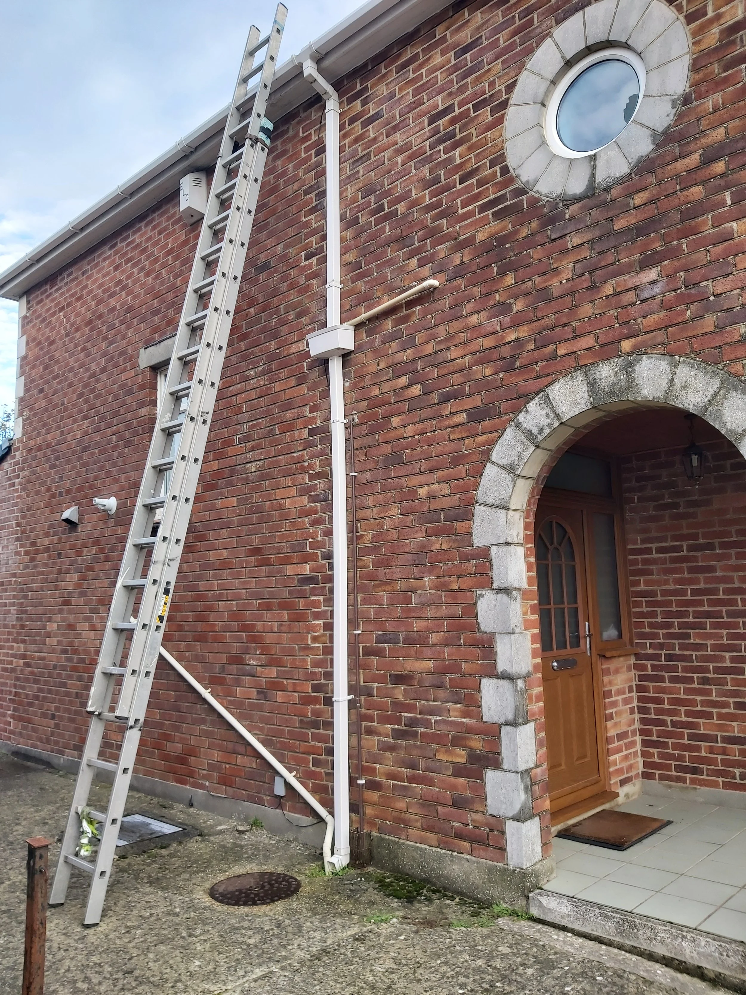 The side of a brick house with a ladder leaning against it, a window near the top, a small white box, a pipe attached to the house, and an arched doorway with a wooden door.