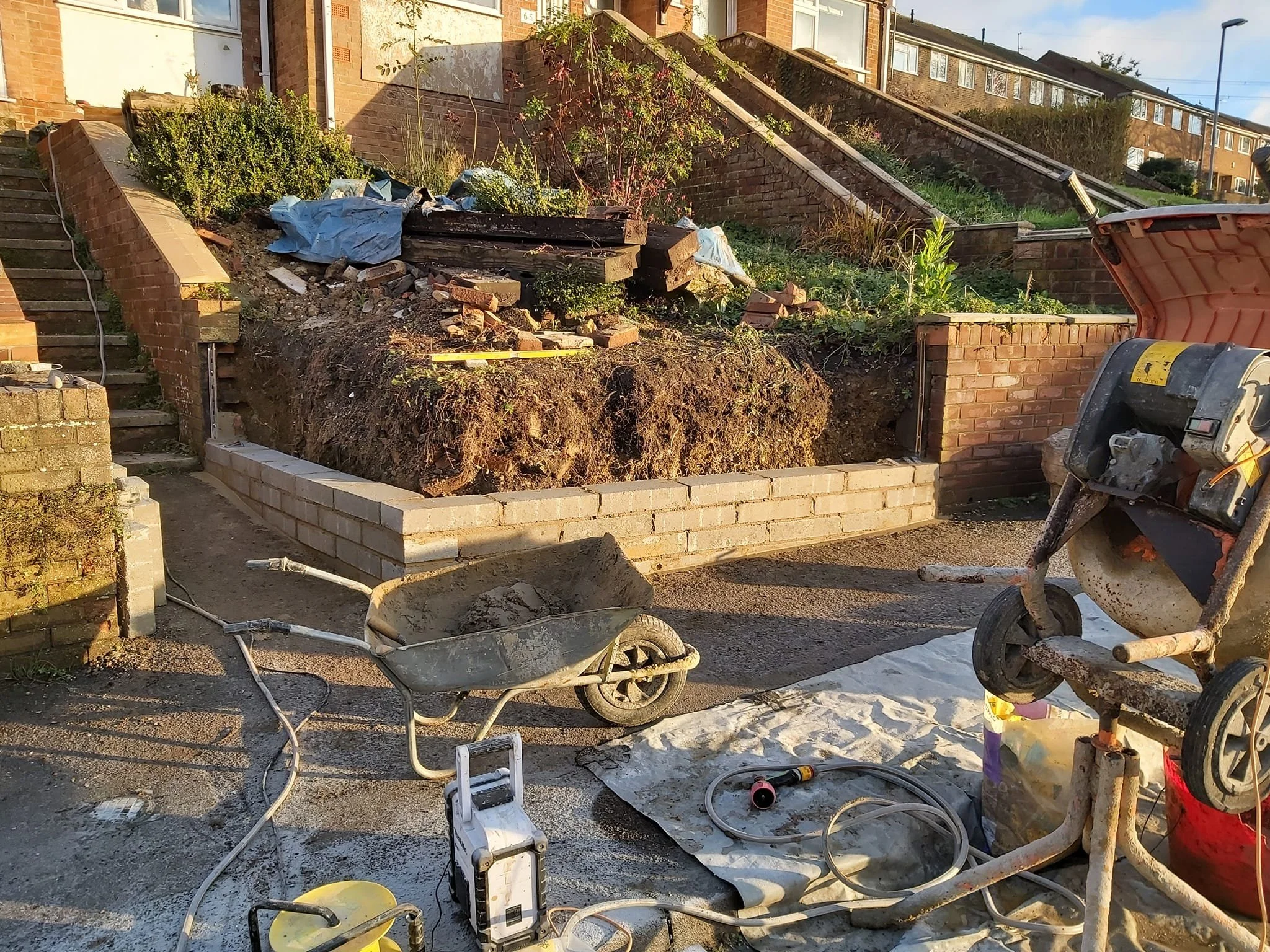 Construction site with a brick wall being built, construction equipment and tools, including a wheelbarrow, a cement mixer, and a white tarp, in front of a residential area with stairs and houses