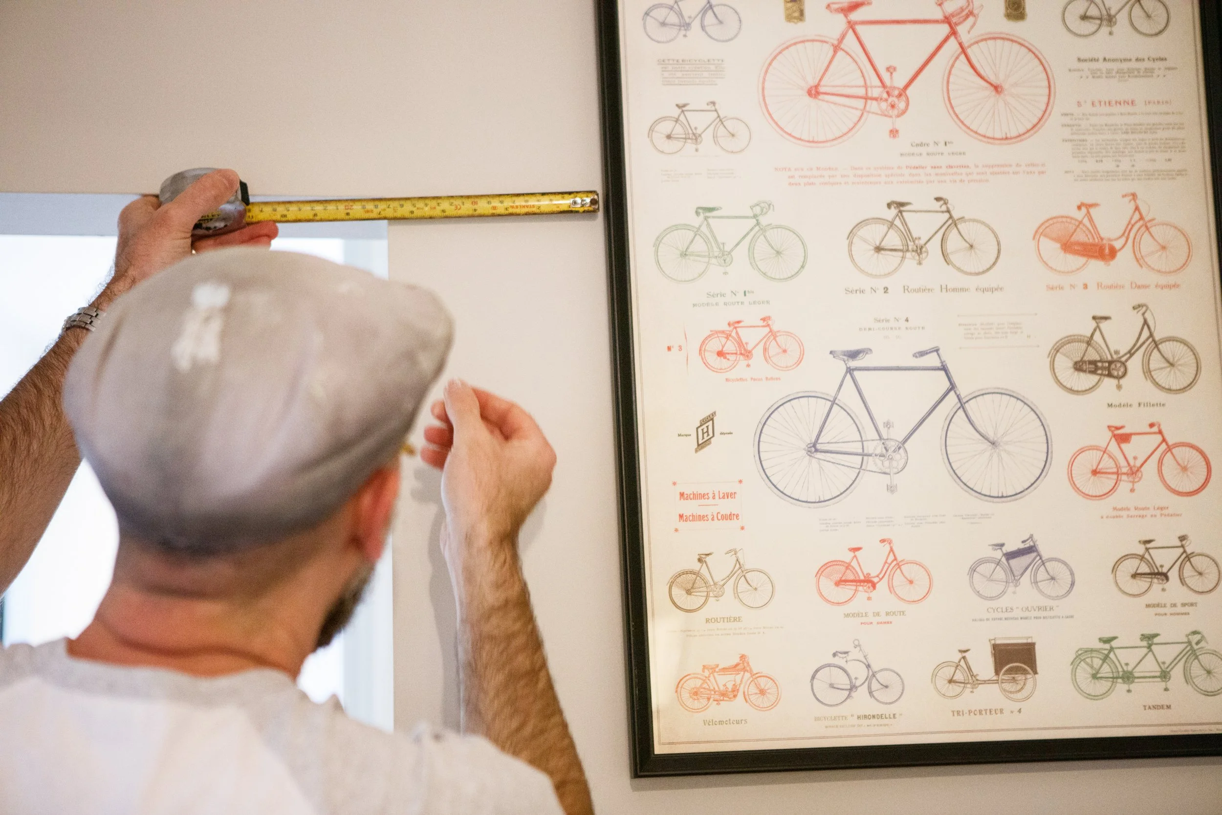 A person wearing a gray beret measures a ceiling section with a yellow tape measure, standing in front of a framed poster displaying different bicycle diagrams and models.